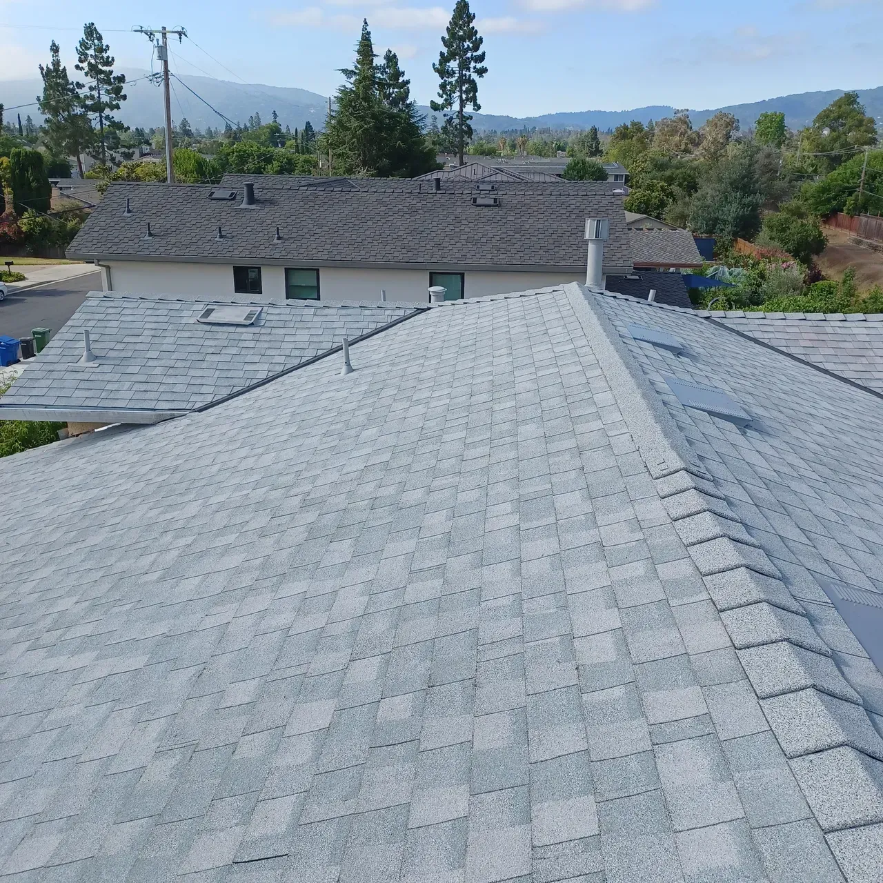 Gray shingle roofs of houses, trees and hills in background under a sunny sky.
