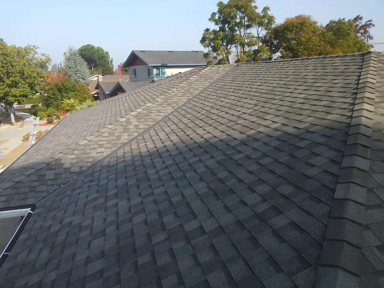 Gray asphalt shingle roof on a house, with other houses and trees in the background under a blue sky.