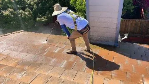 Person in safety gear power washing a brown shingle roof.