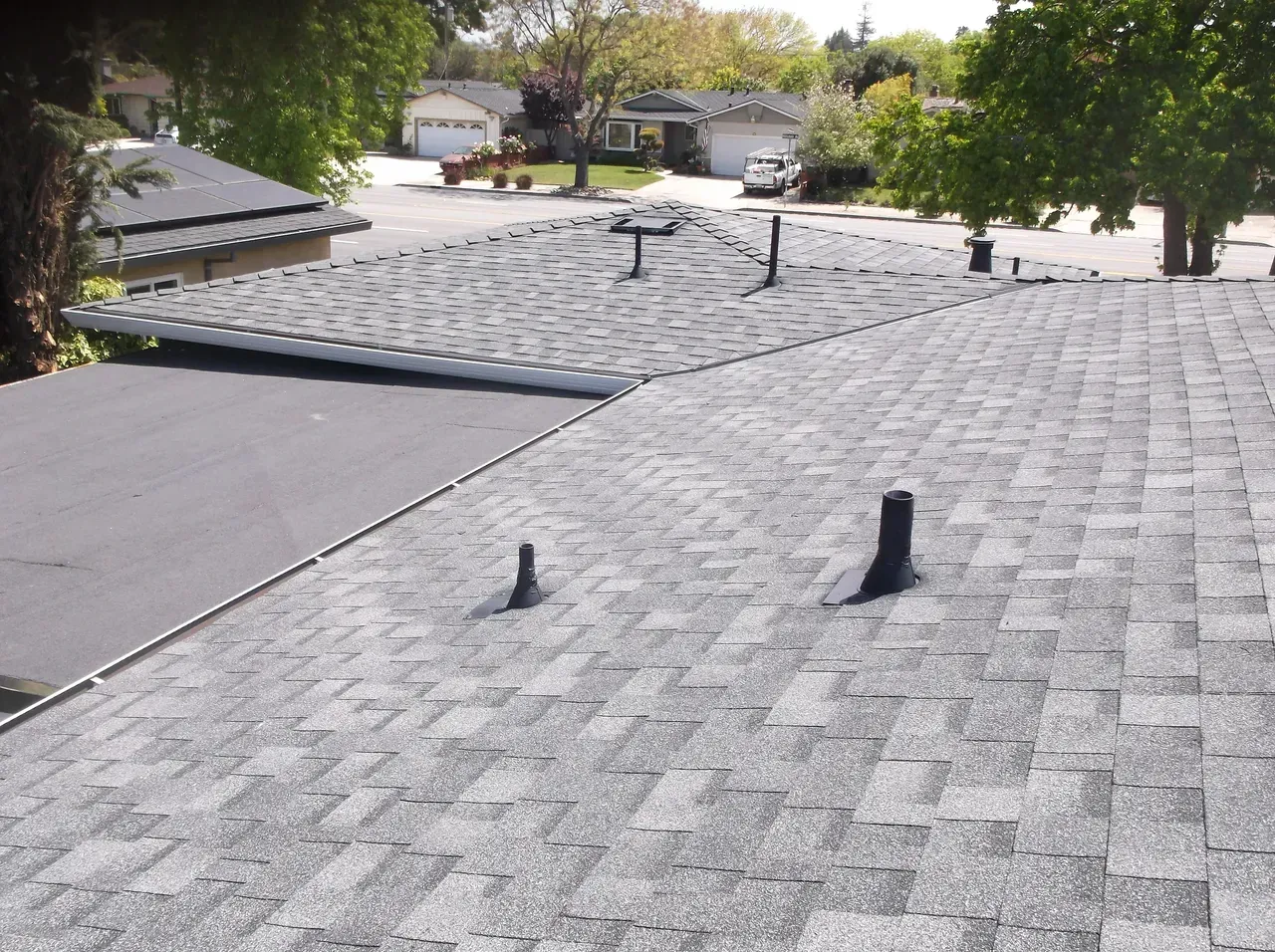 A rooftop with grey asphalt shingles and black vents, overlooking a residential neighborhood.