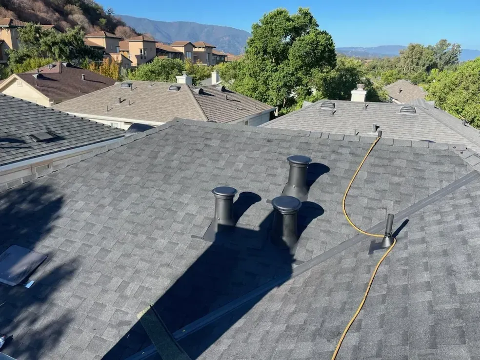 View of dark gray shingled rooftops, vent pipes, and a yellow cord under a bright blue sky.