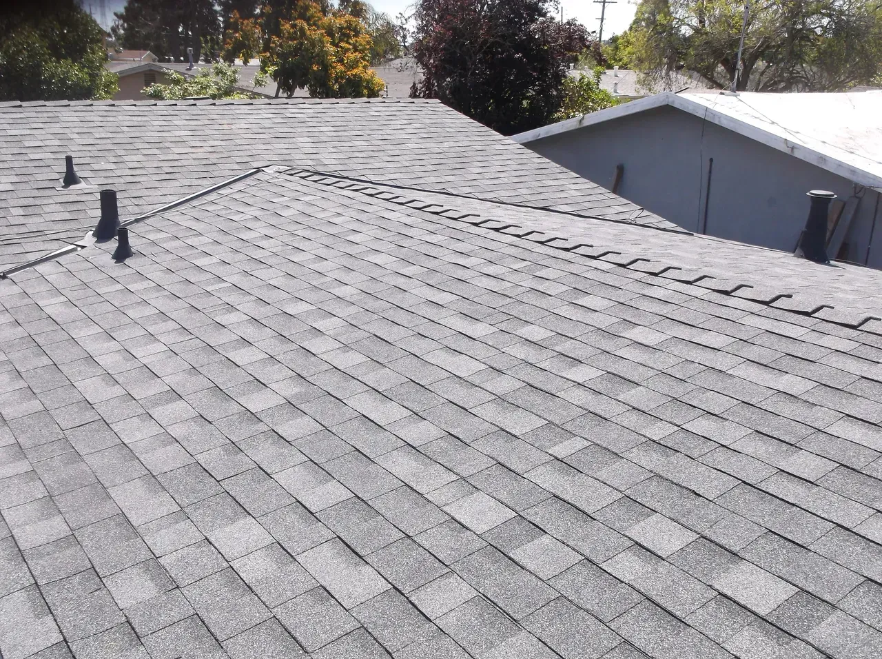 Gray asphalt shingle roof on a house, with pipes and another roof visible in the background.