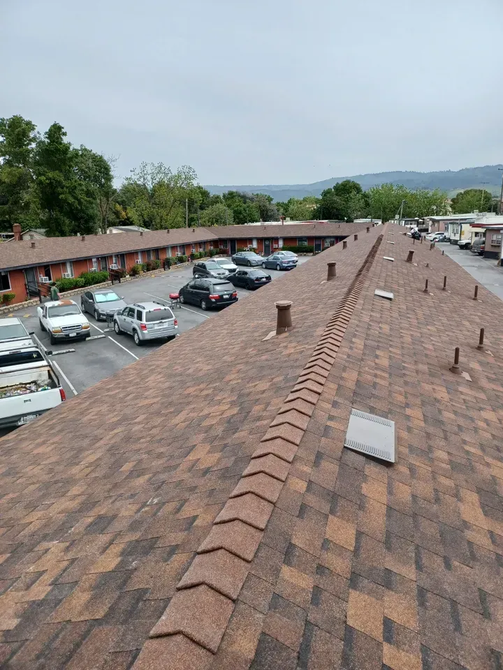 Rooftop view of a motel with parked cars in the lot. Brown shingle roof, cloudy sky.