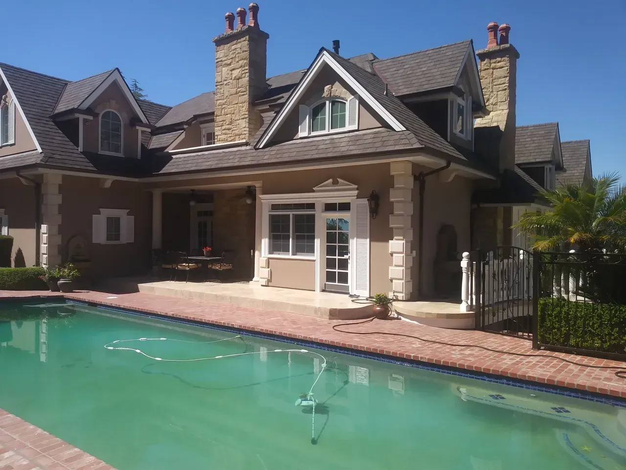 House with pool: Tan stucco, stone trim, dark roof, two chimneys, arched window, and red brick pool surround.
