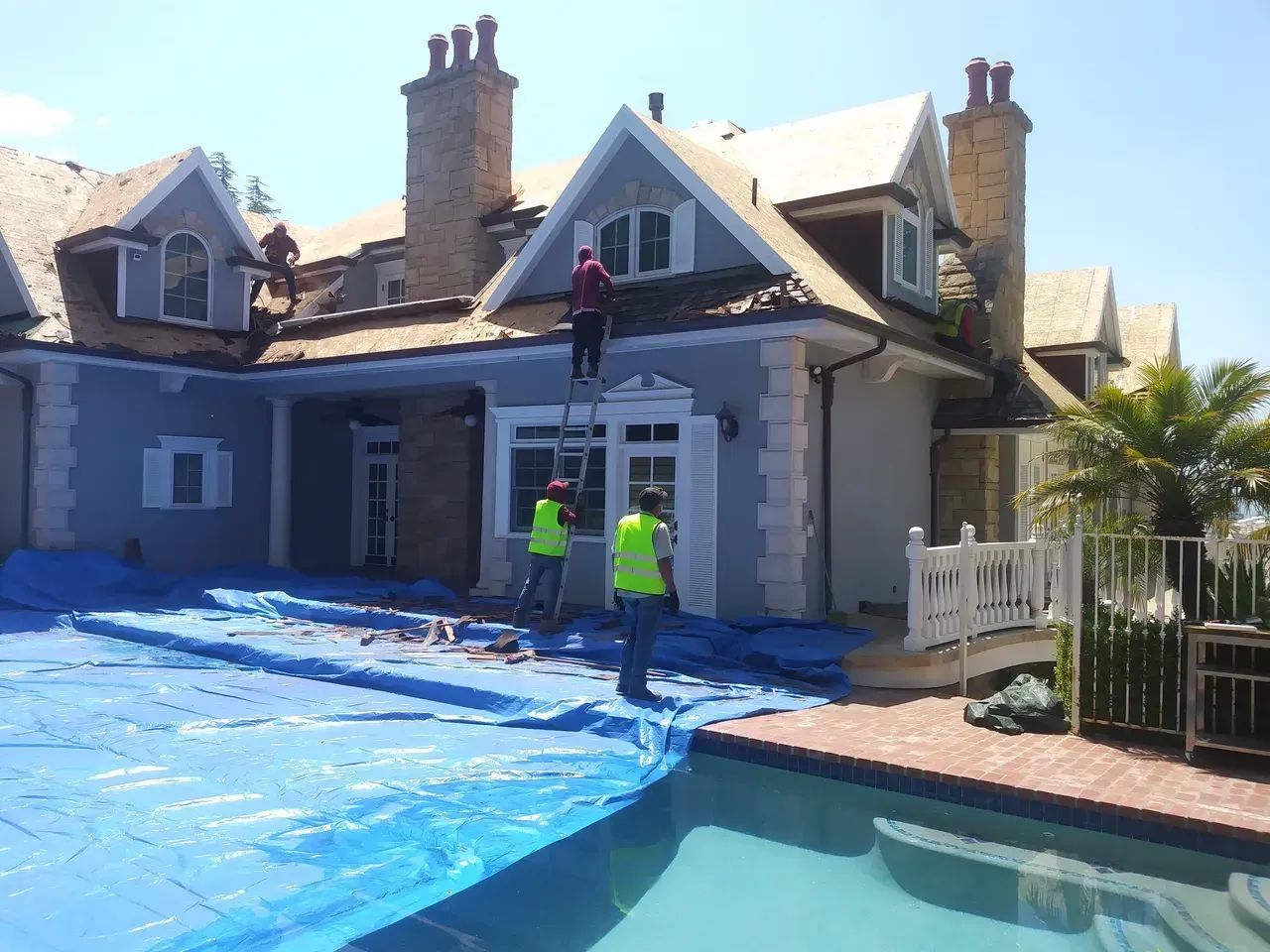 Workers on ladder, repairing a house roof, blue tarp covers the ground, pool in foreground.