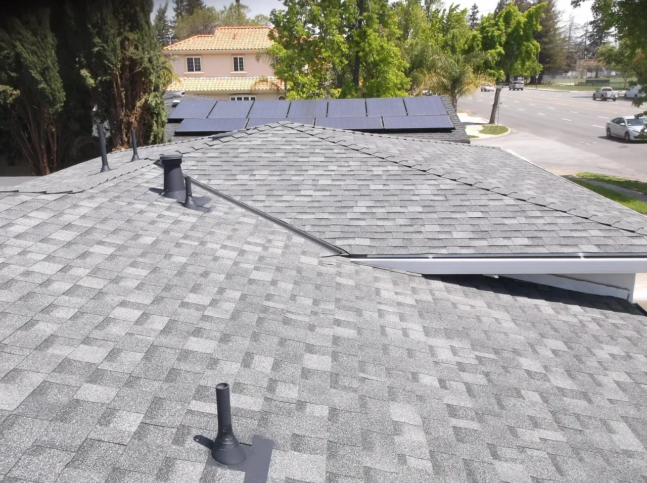 Gray shingled roof with solar panels, vent pipes, and a house in the background. Sunny day.
