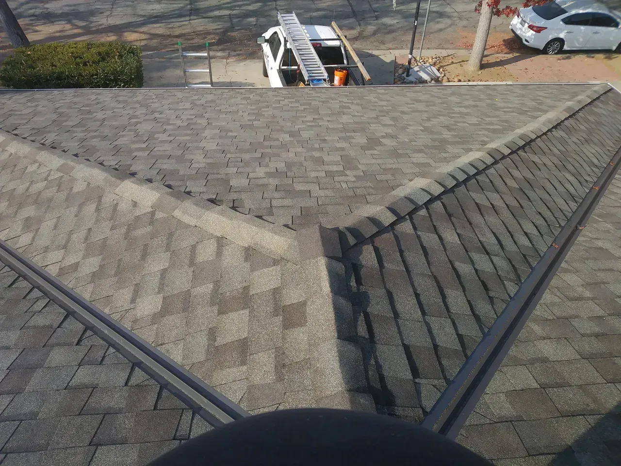 Overhead view of a roof with gray shingles, a metal ridge cap, and a work van parked below.