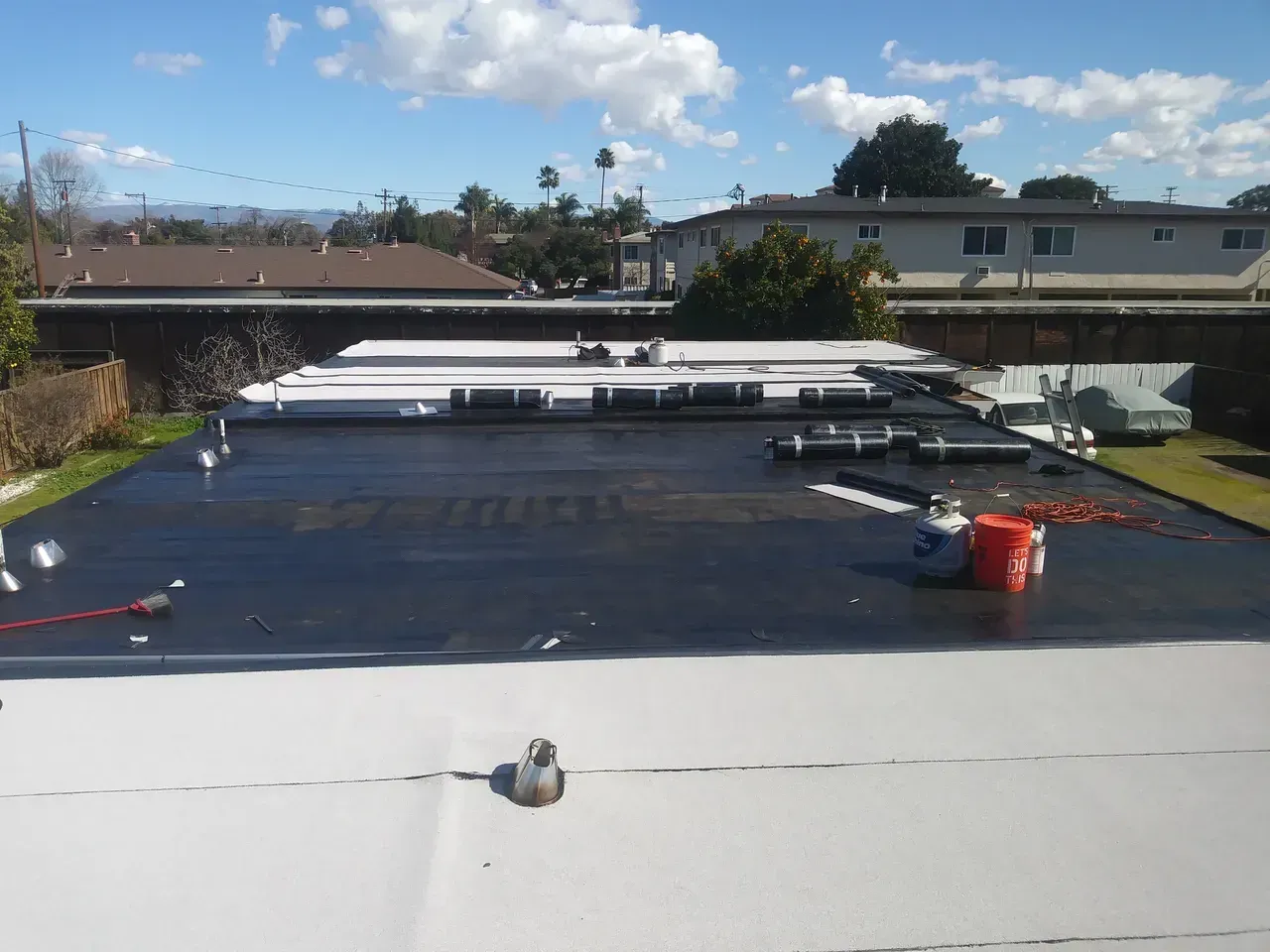 Flat roof with patches of black tar and white coating, equipment and buckets visible. Sunny day.