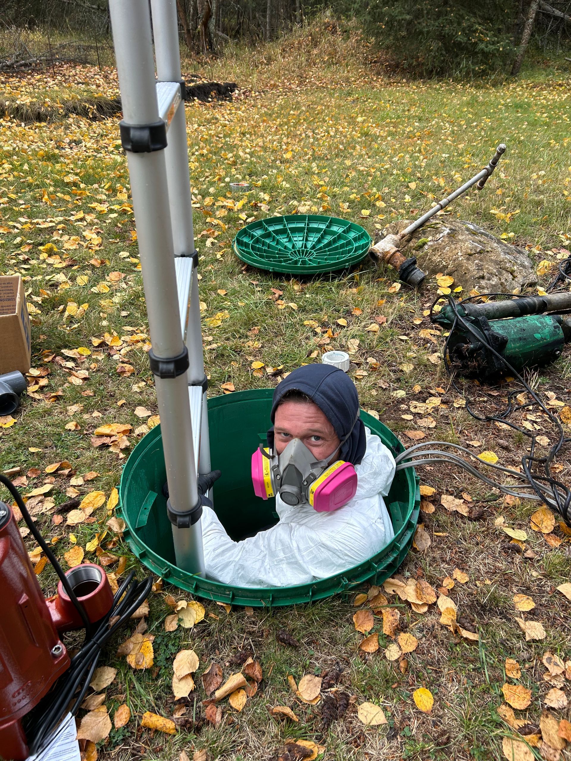 Person in protective suit and respirator mask inside a septic tank, near ladder and open hatch outdoors.