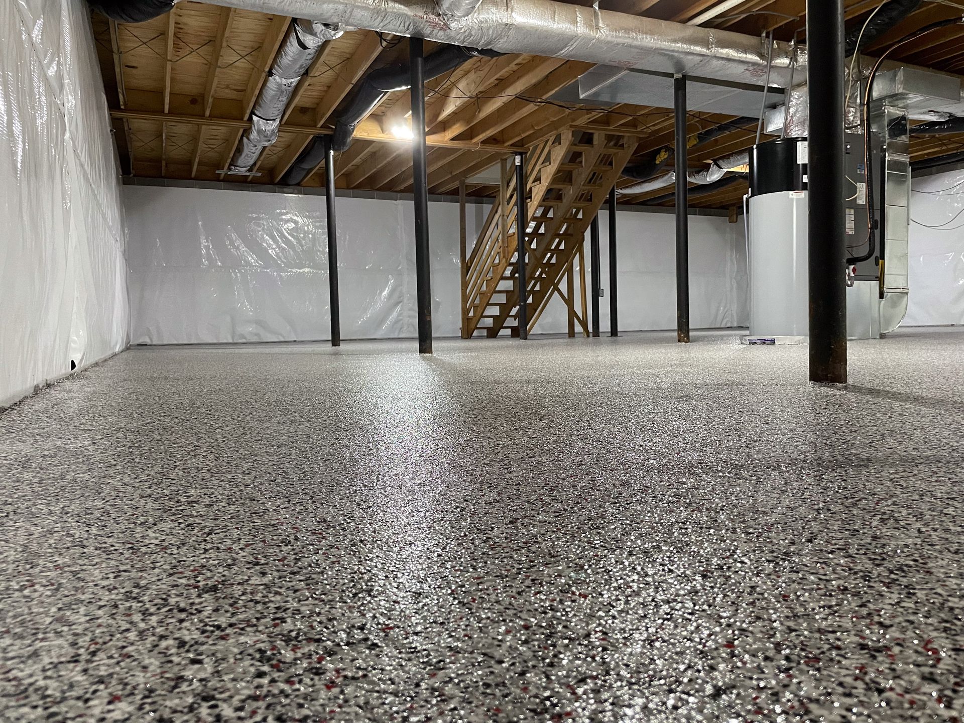 A finished basement with a glossy, multi-colored speckled epoxy floor, white vapor barrier walls, and wooden stairs.