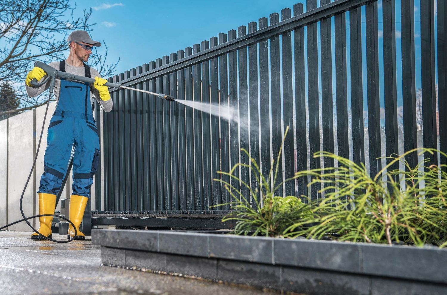 Person power washing a dark metal fence, wearing blue overalls, yellow boots and gloves, outdoors.