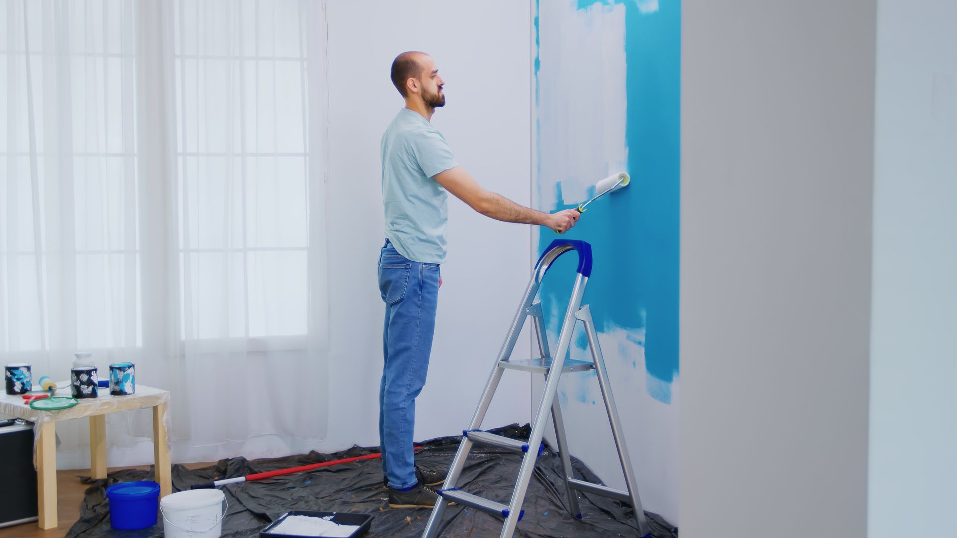 A man is standing on a ladder painting a wall blue.
