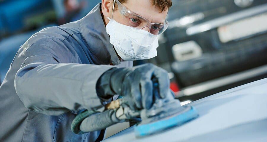 A person wearing safety glasses, a mask, and gloves sands a car's surface with a power sander in a repair shop.