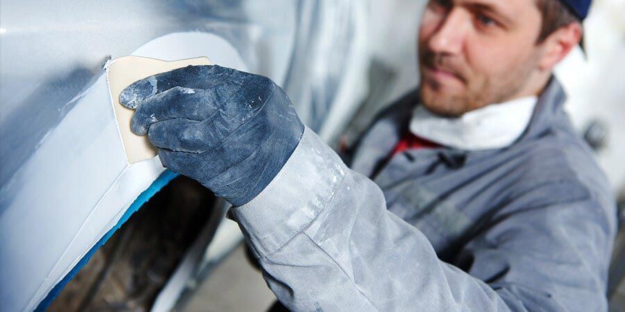 A mechanic in a blue jumpsuit sands a car panel while wearing a black glove.