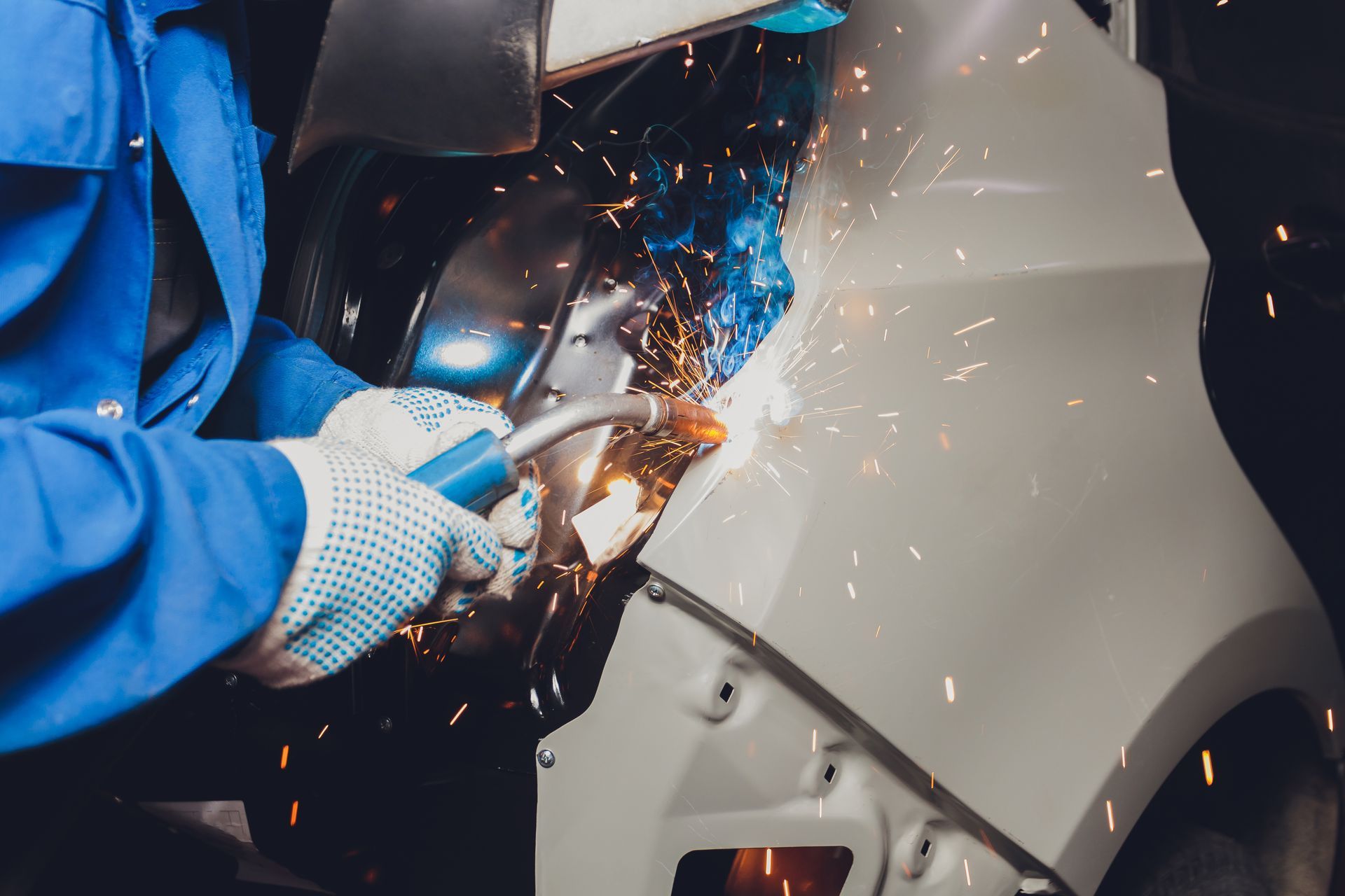 A worker in a blue uniform uses a welding tool on a car panel, creating bright sparks.