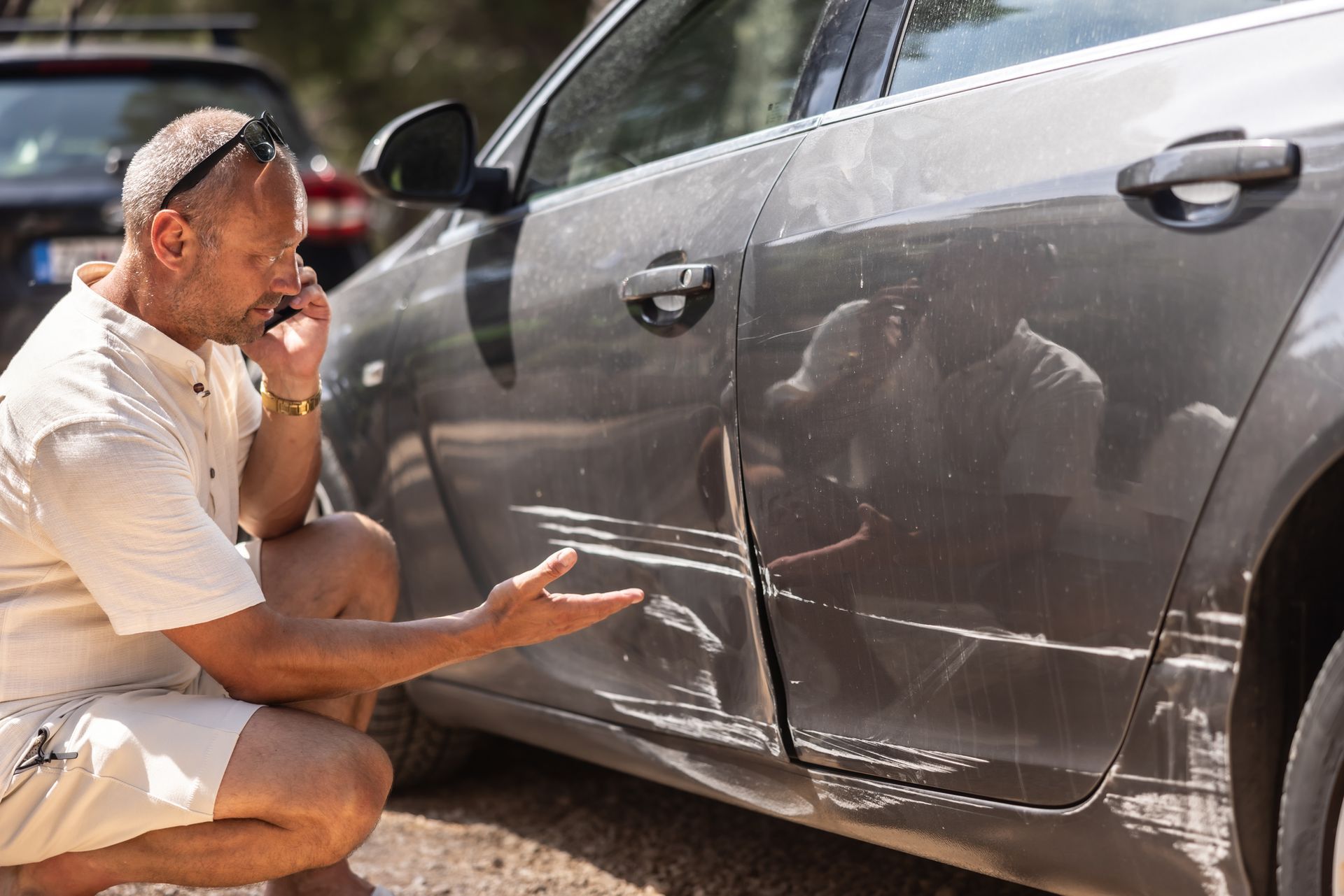 A man, kneeling in front of a car with a damaged door, makes a call, under the sun.