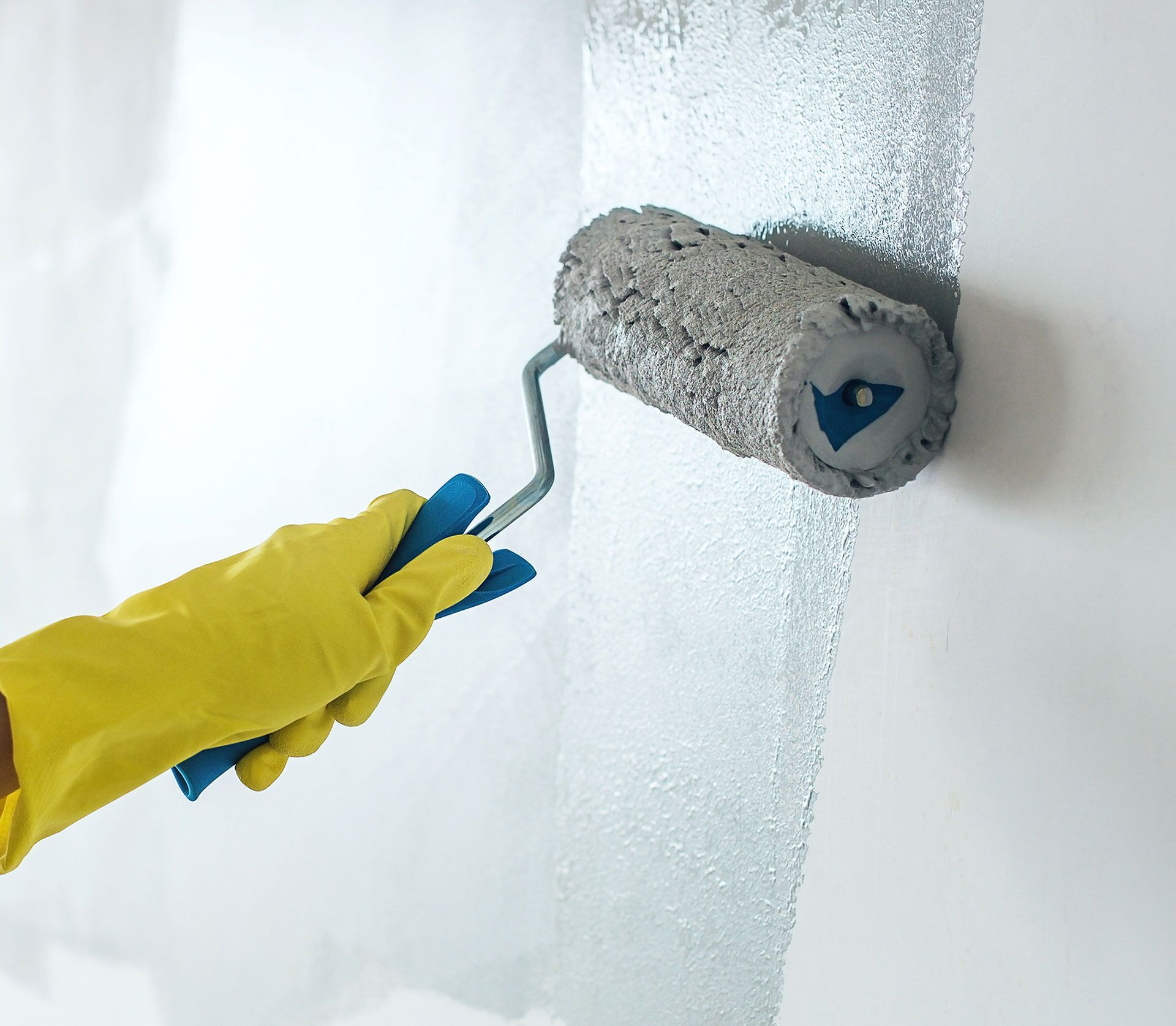 Person in yellow glove painting a white wall with a paint roller.
