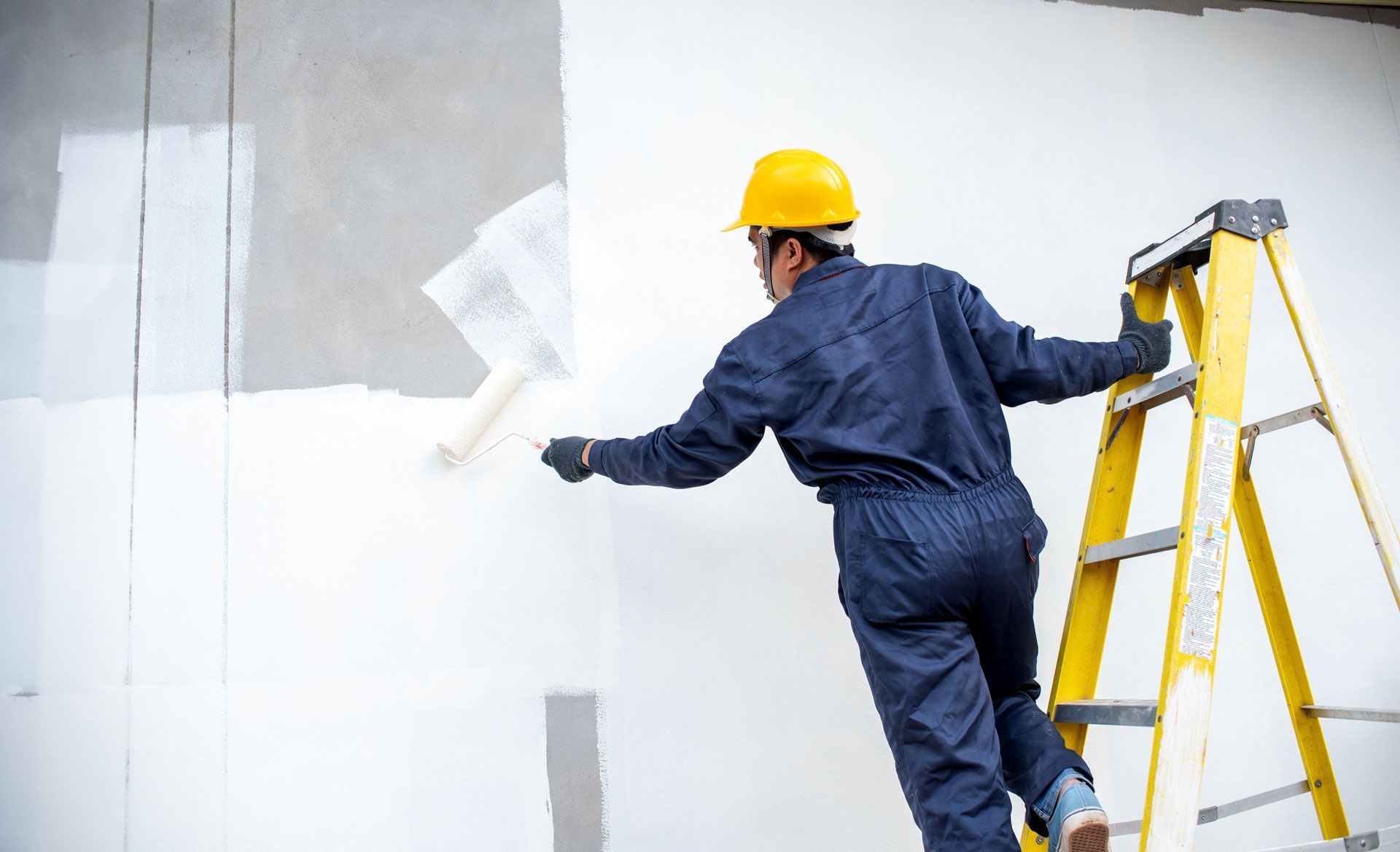 Person in blue jumpsuit and yellow hardhat painting a wall white, using a roller, while standing on a yellow ladder.
