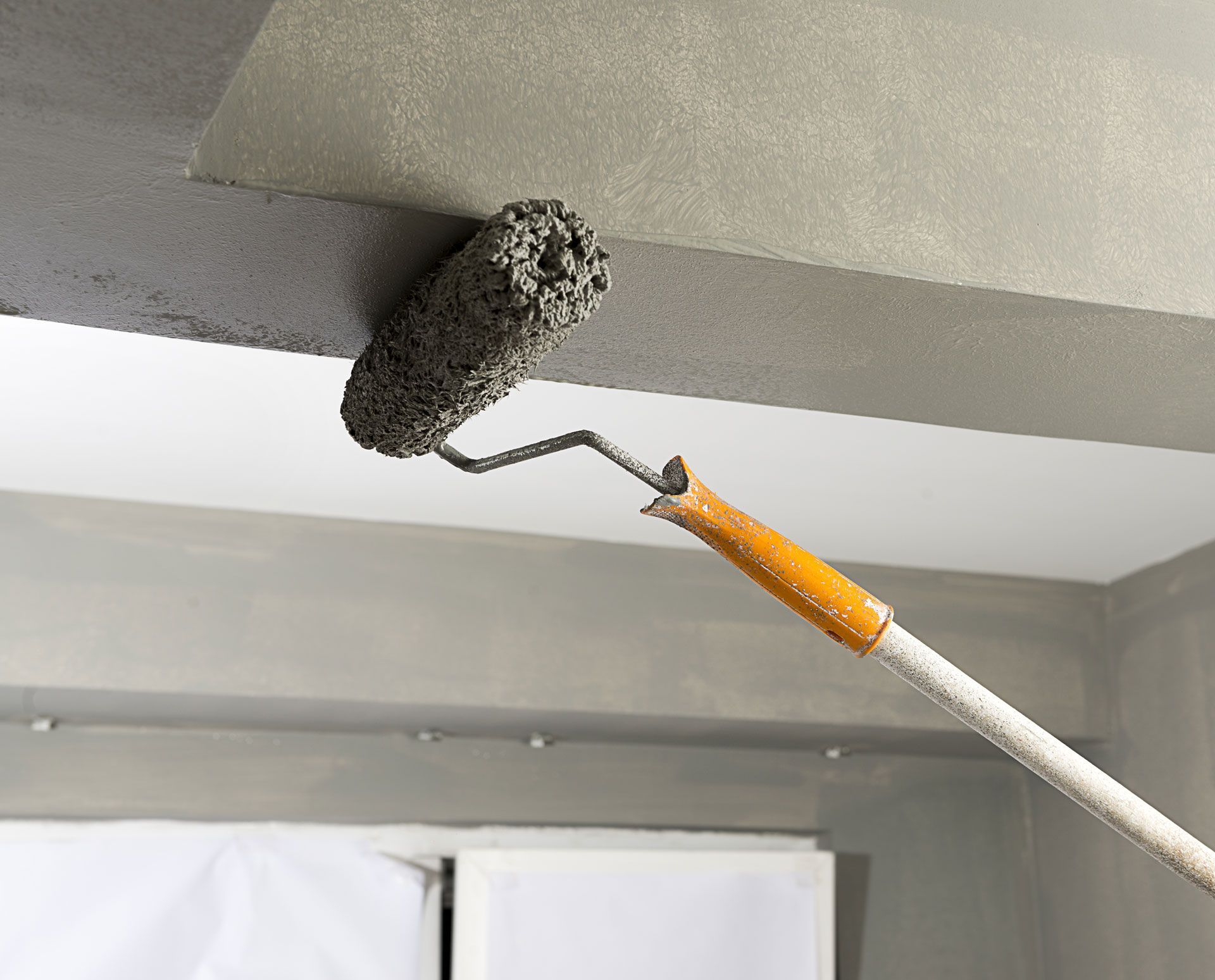 A paint roller with a textured head is used to paint a gray beam in a room.