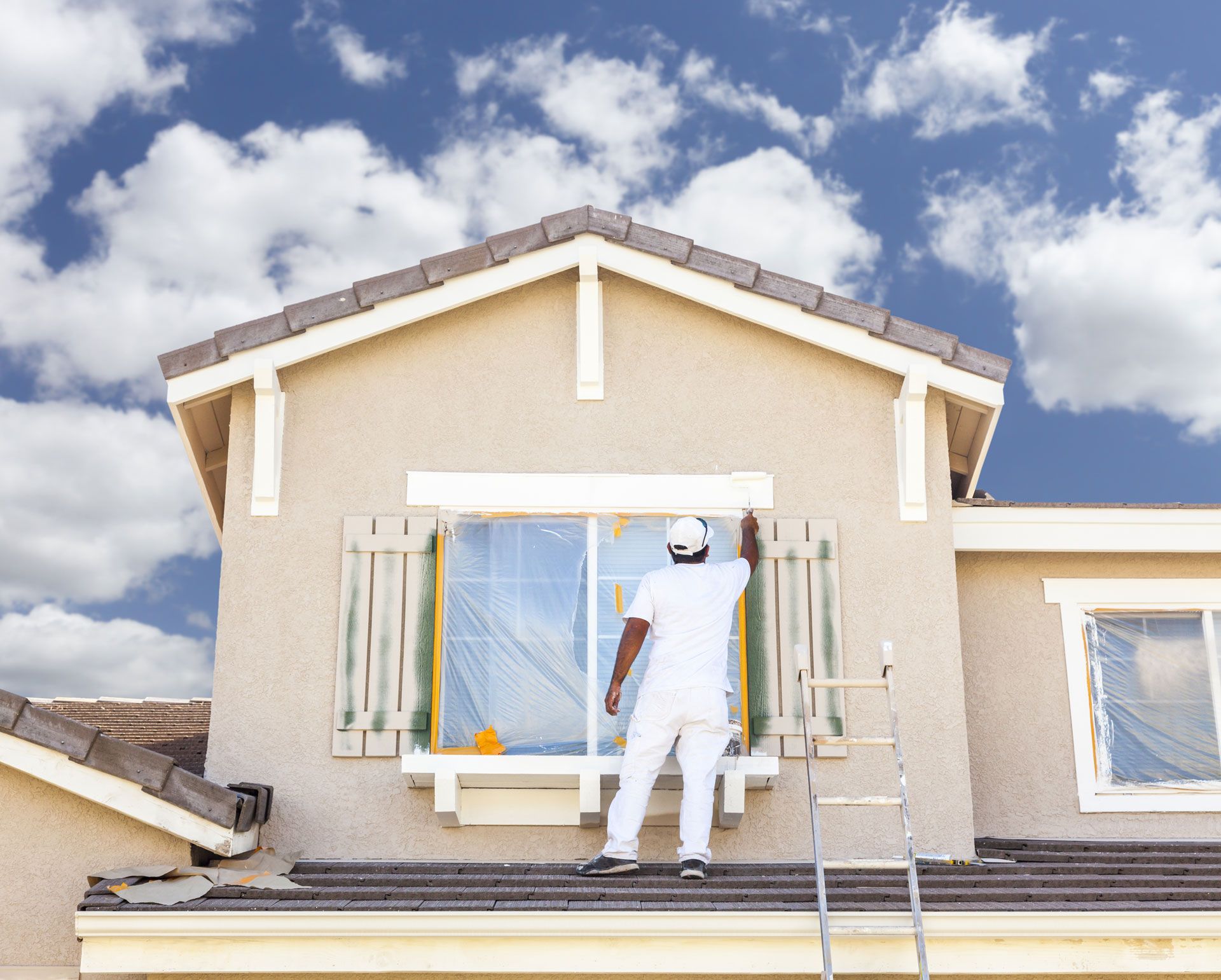 A person in white coveralls painting the exterior trim of a two-story beige house under a cloudy sky.