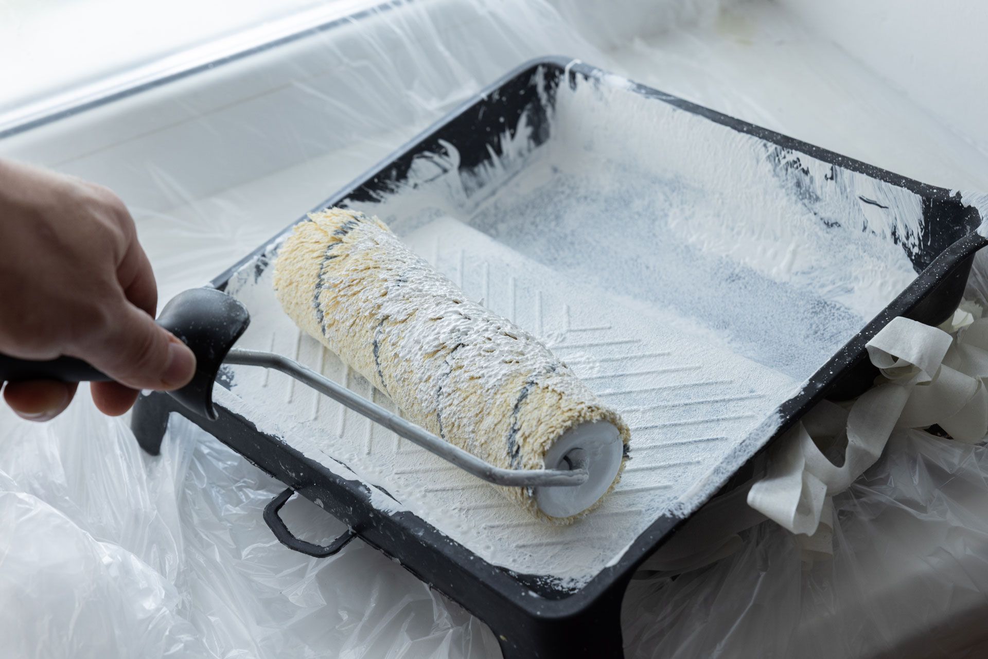 Hand holding paint roller in tray filled with white paint, next to a window.