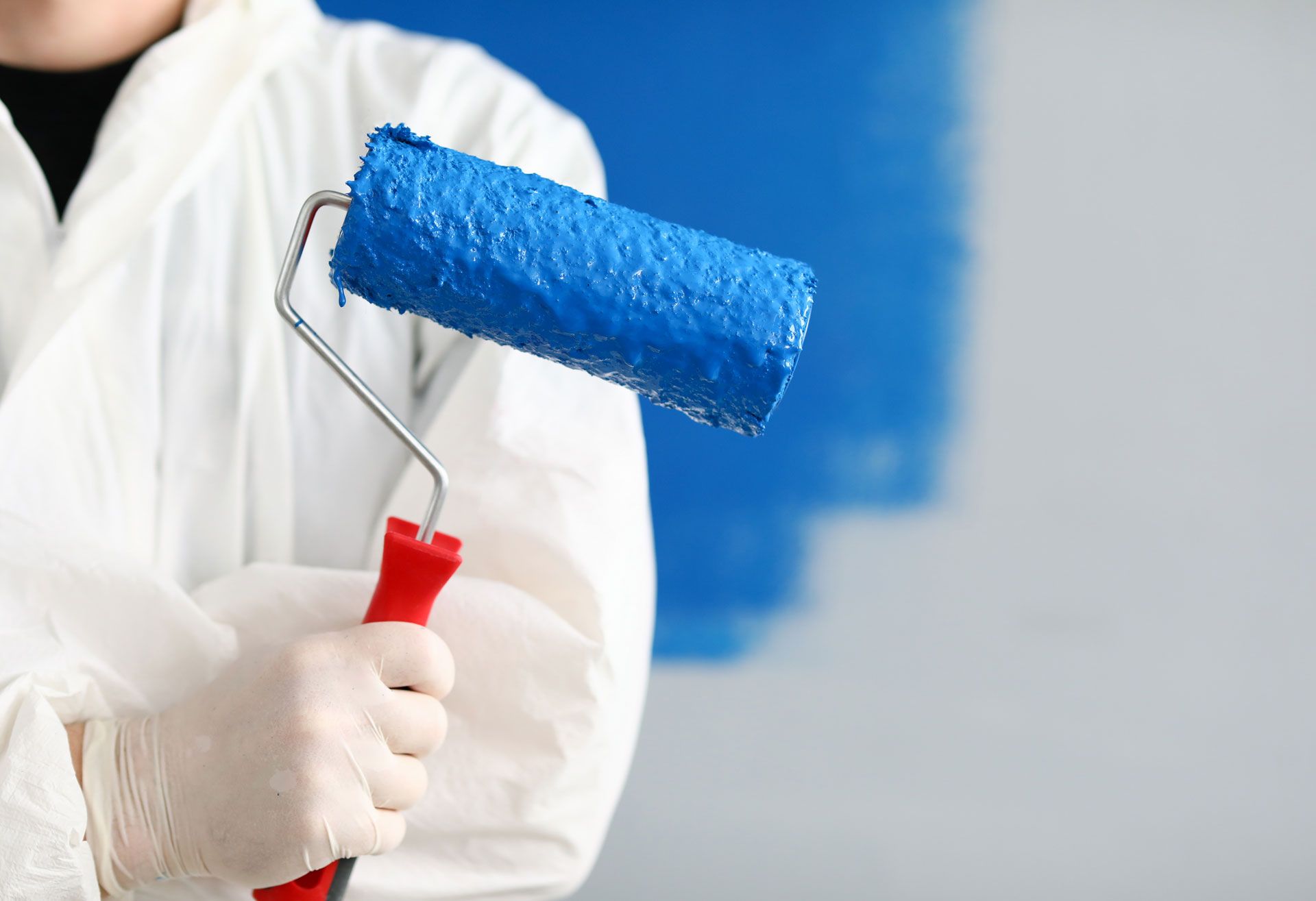 Person in white protective suit holds a blue paint roller, painting a wall blue.