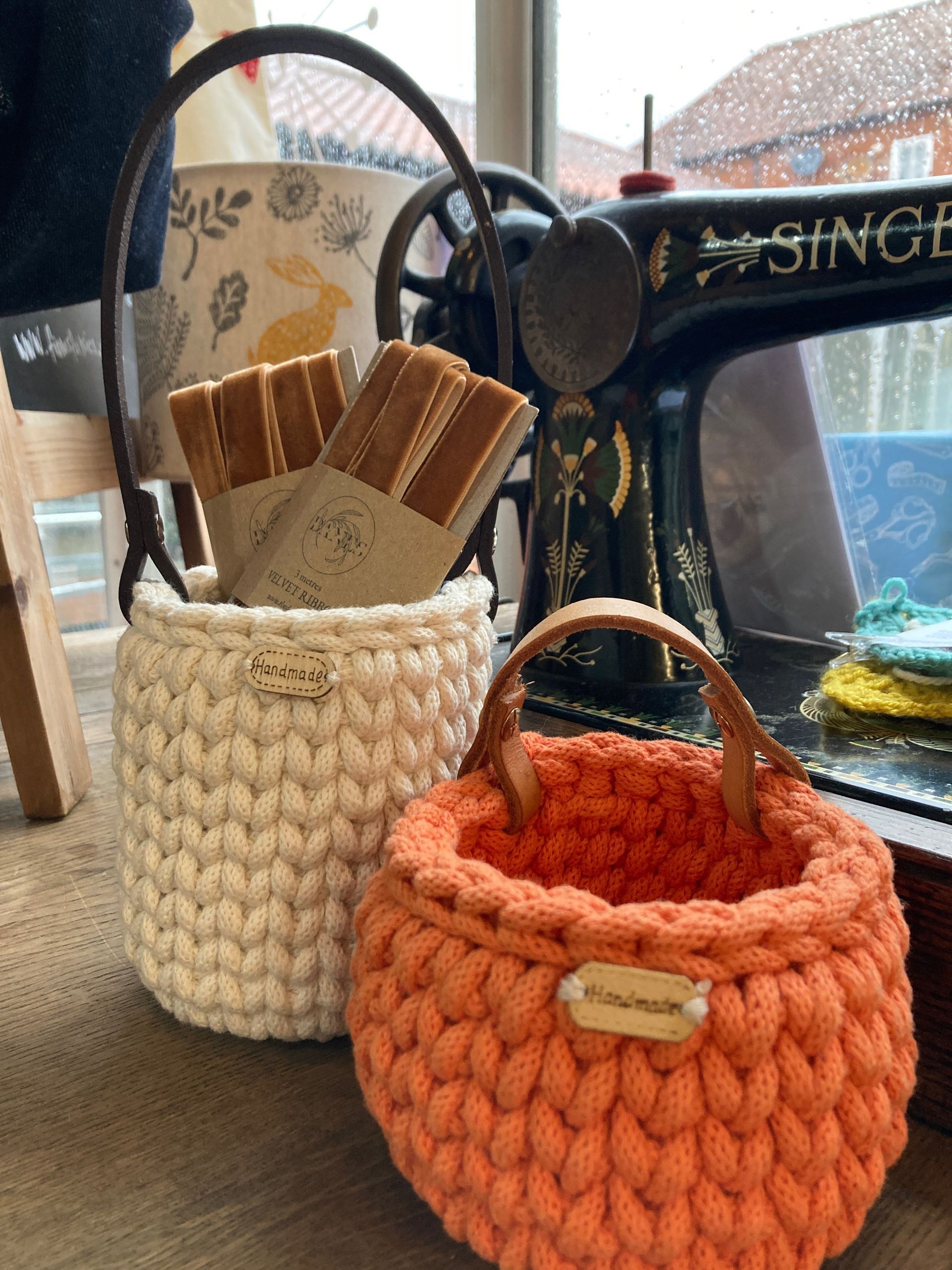 Two handmade crochet baskets, one cream, one orange, with leather handles, near a sewing machine.