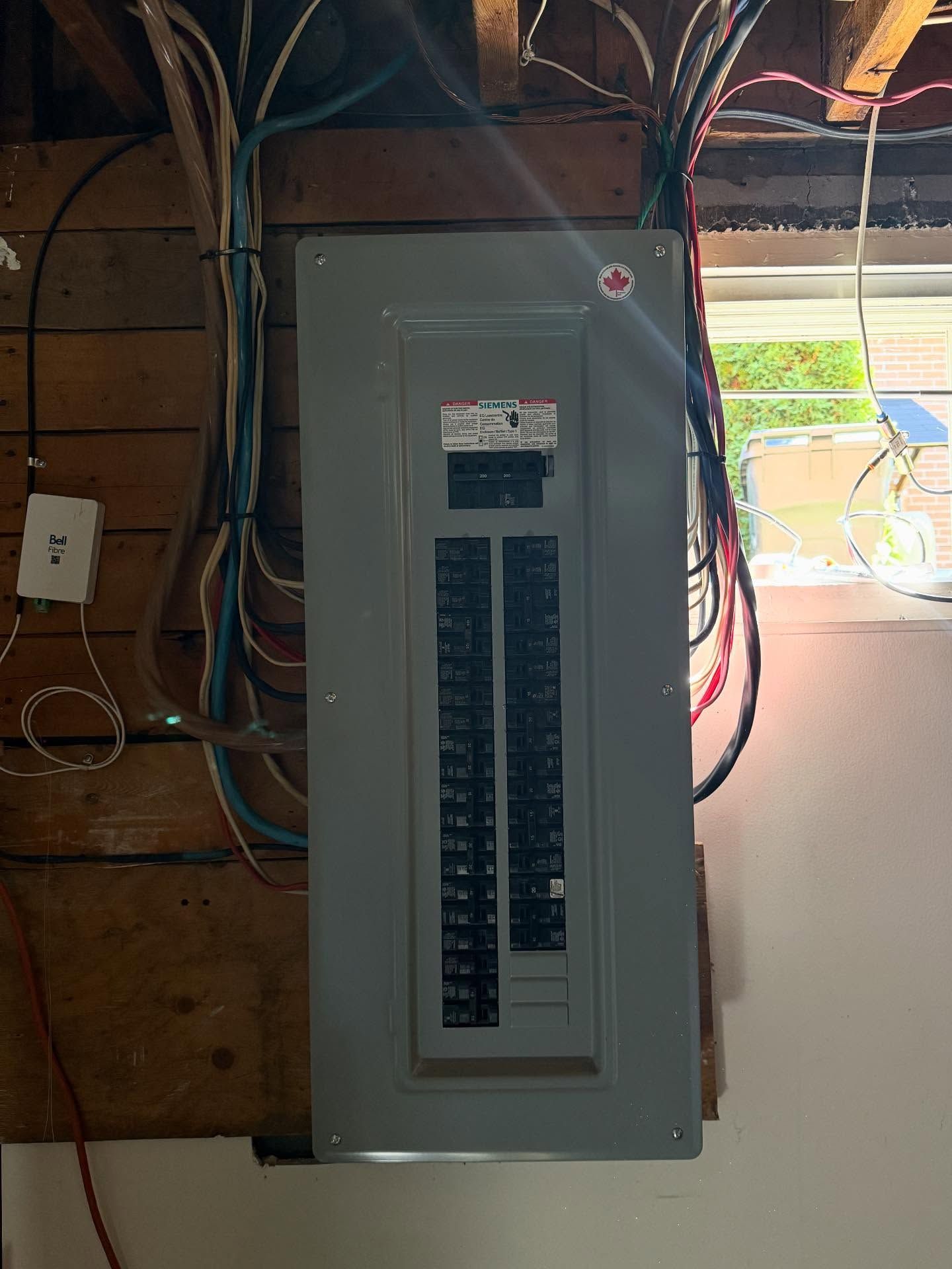 A grey electrical breaker panel mounted on a wood-slatted wall in a room with a window to the right.