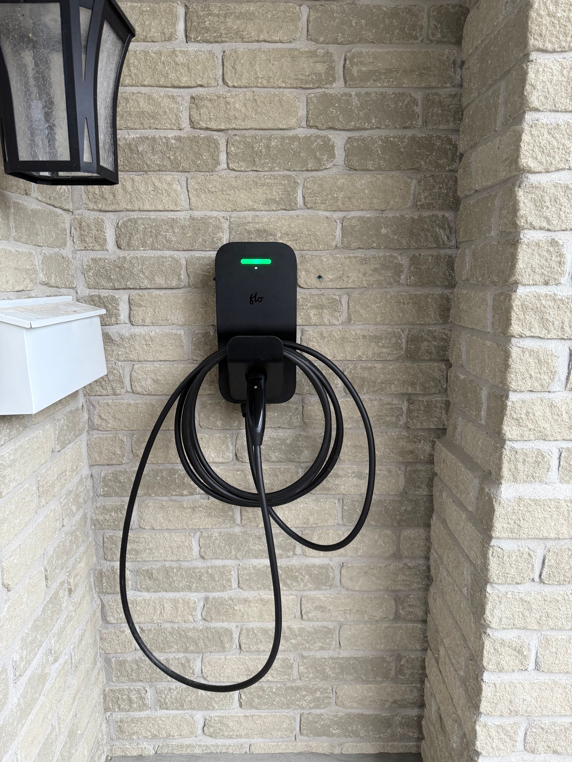 A black EV charging station mounted on a beige brick wall with a coiled black cable hanging from it.