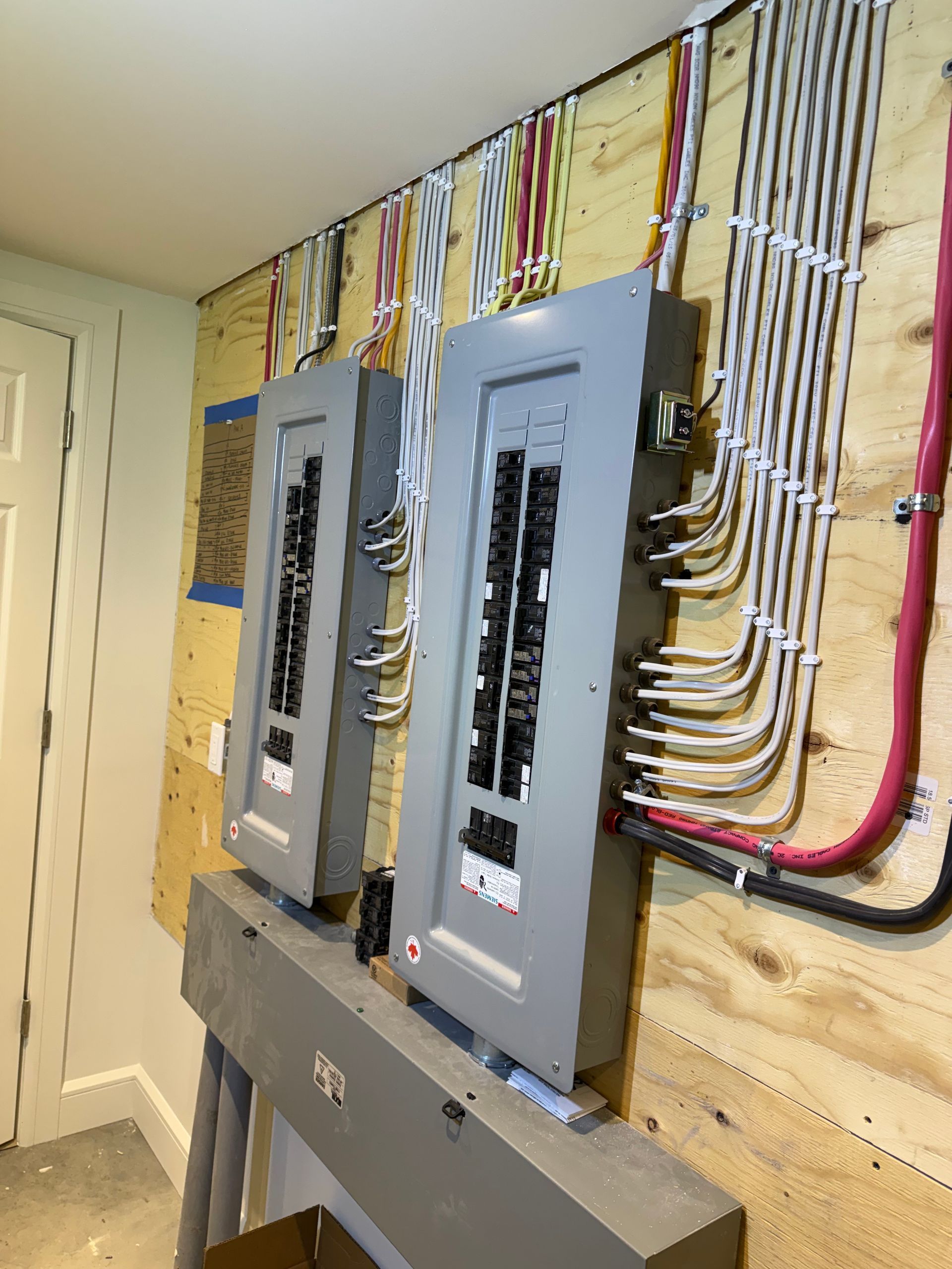 Two grey electrical breaker panels mounted on a plywood wall with numerous white and colored wires routed neatly.