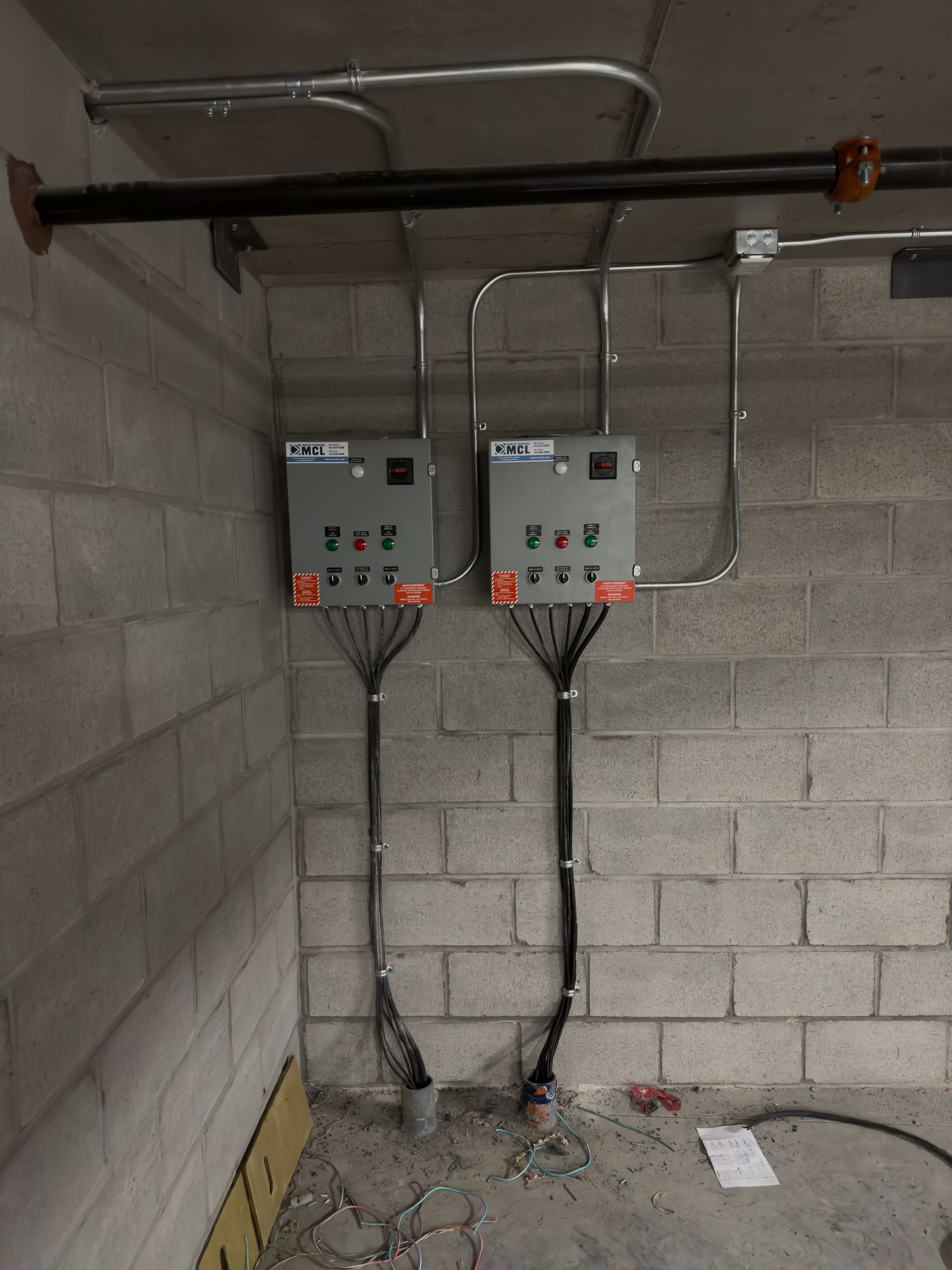 Deux boîtiers de commande électriques gris industriel, montés sur un mur en parpaings, avec tuyauterie métallique et câblage noir.