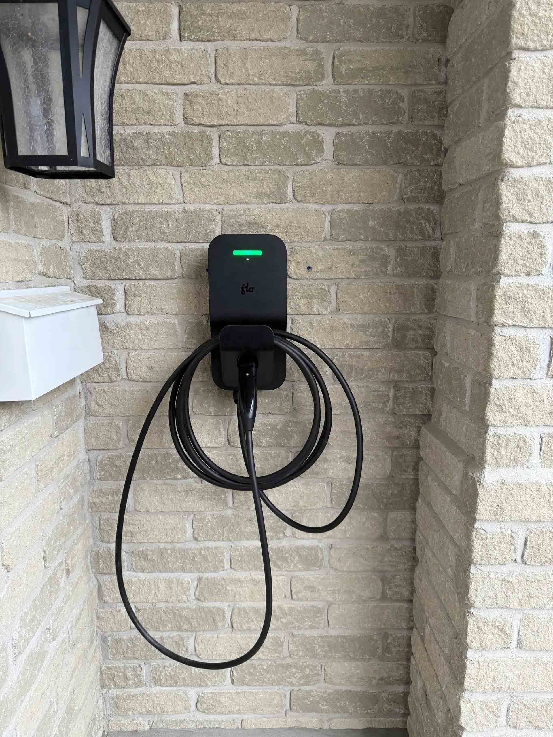 Une borne de recharge pour véhicules électriques avec un câble noir enroulé, fixée sur un mur de briques claires.