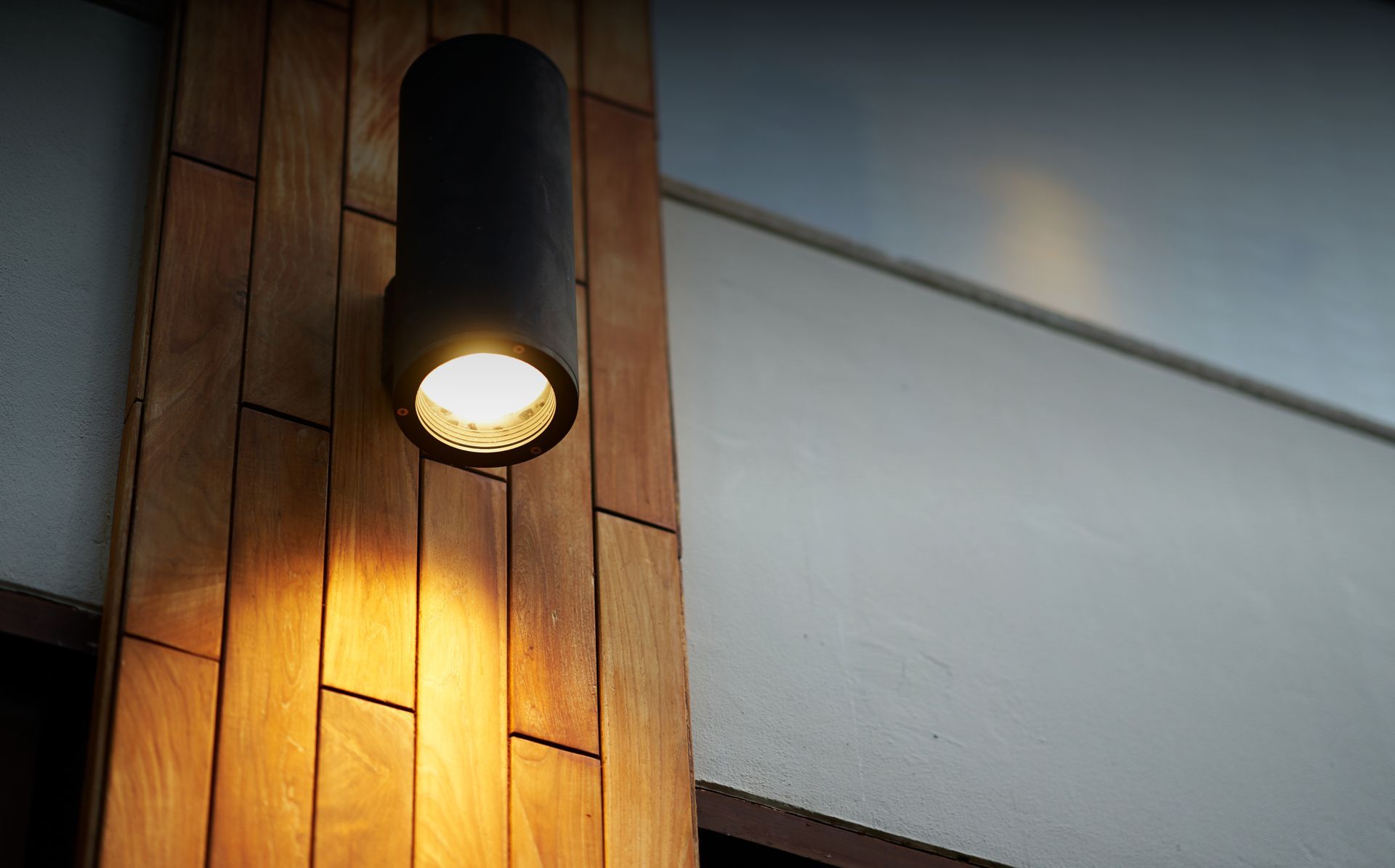 A cylindrical outdoor wall lamp emits a warm, downward glow, mounted on a vertical wooden plank feature wall.