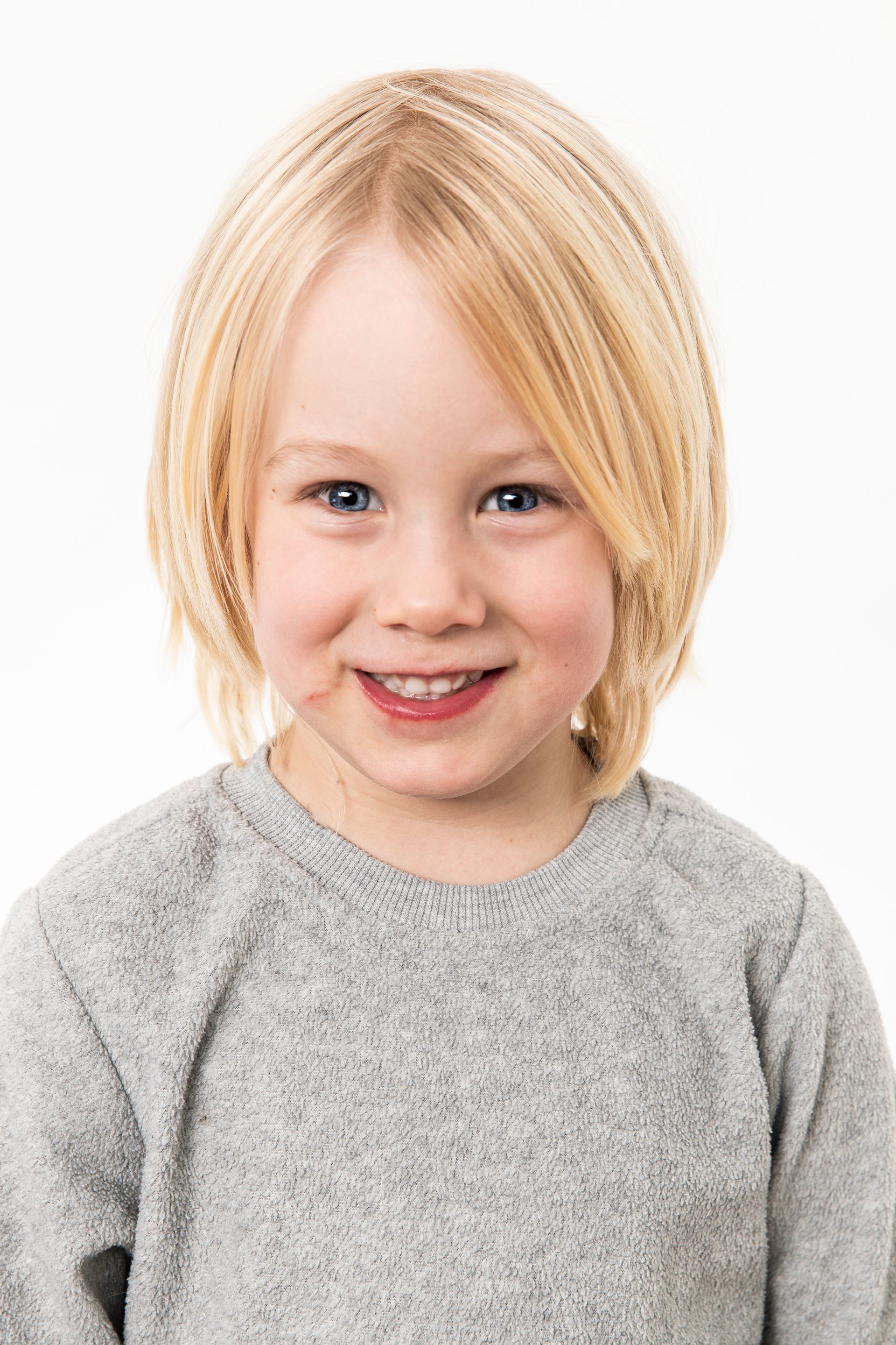 Een klein jongetje met blond haar draagt ​​een grijze trui en glimlacht.