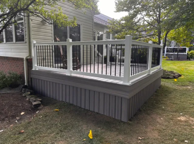 Raised gray deck with white railing attached to a house, surrounded by a yard and trees