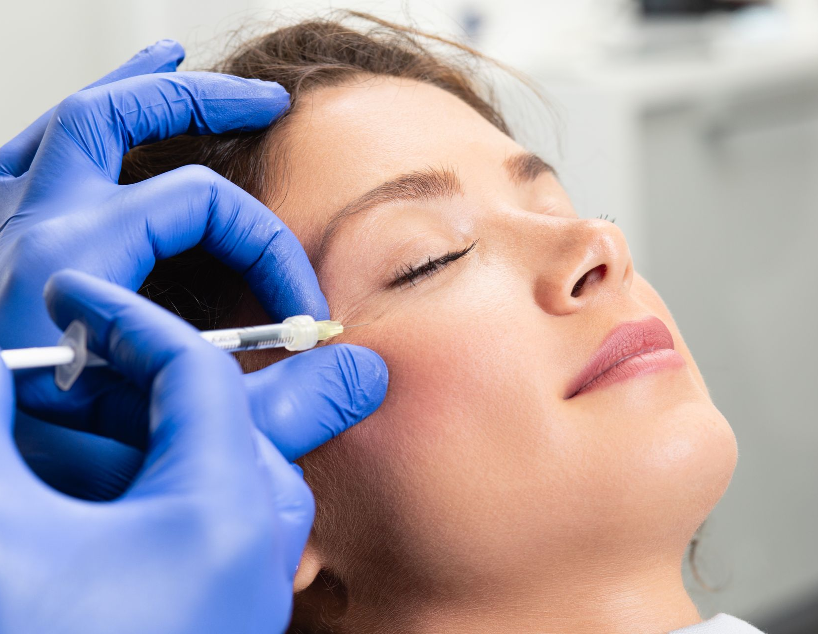 Person receiving facial injections, doctor's gloved hand holding syringe near eye.