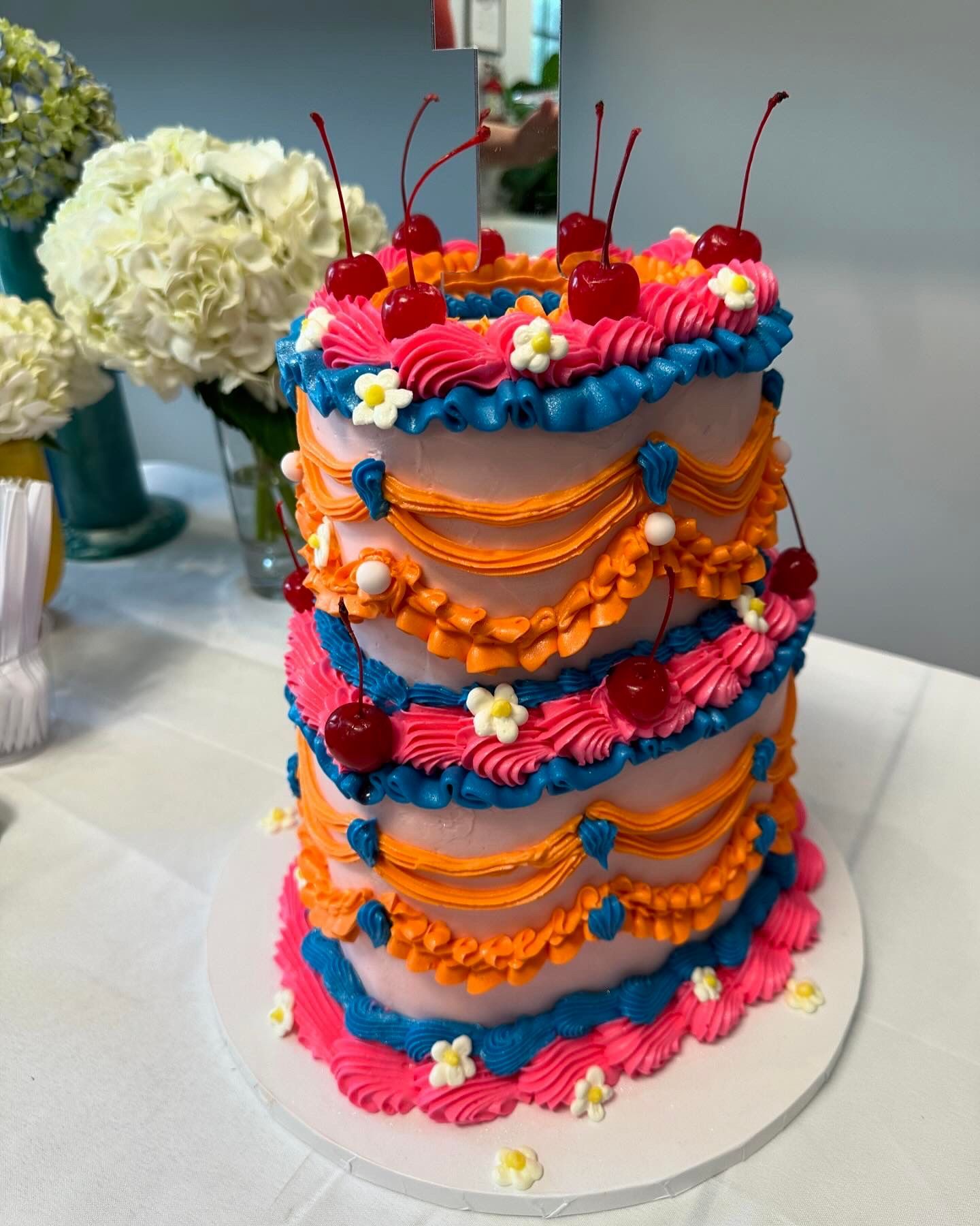 Layered cake decorated with pink, orange, blue frosting, cherries, and flowers.