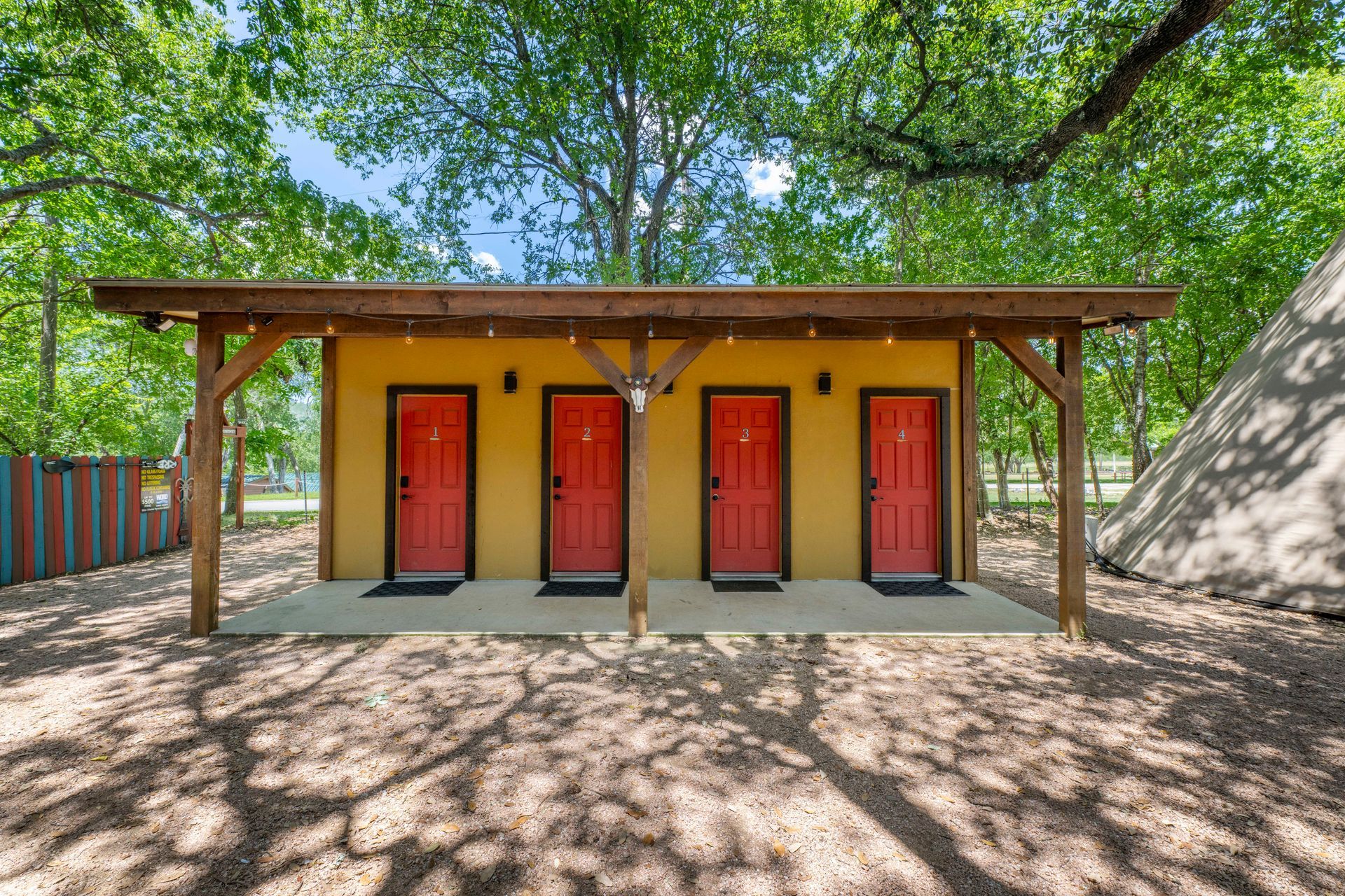 Private bath house four red doors on a yellow building under a wooden awning, trees overhead, and a teepee to the right.