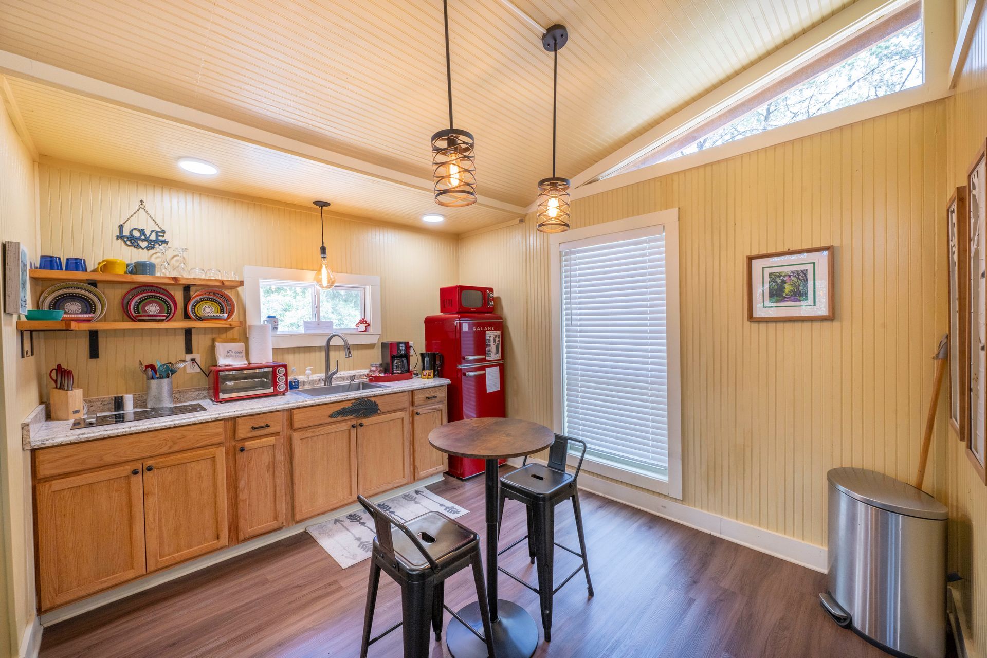 Cozy yellow kitchen with wooden cabinets, red fridge, round table, and hanging lights.