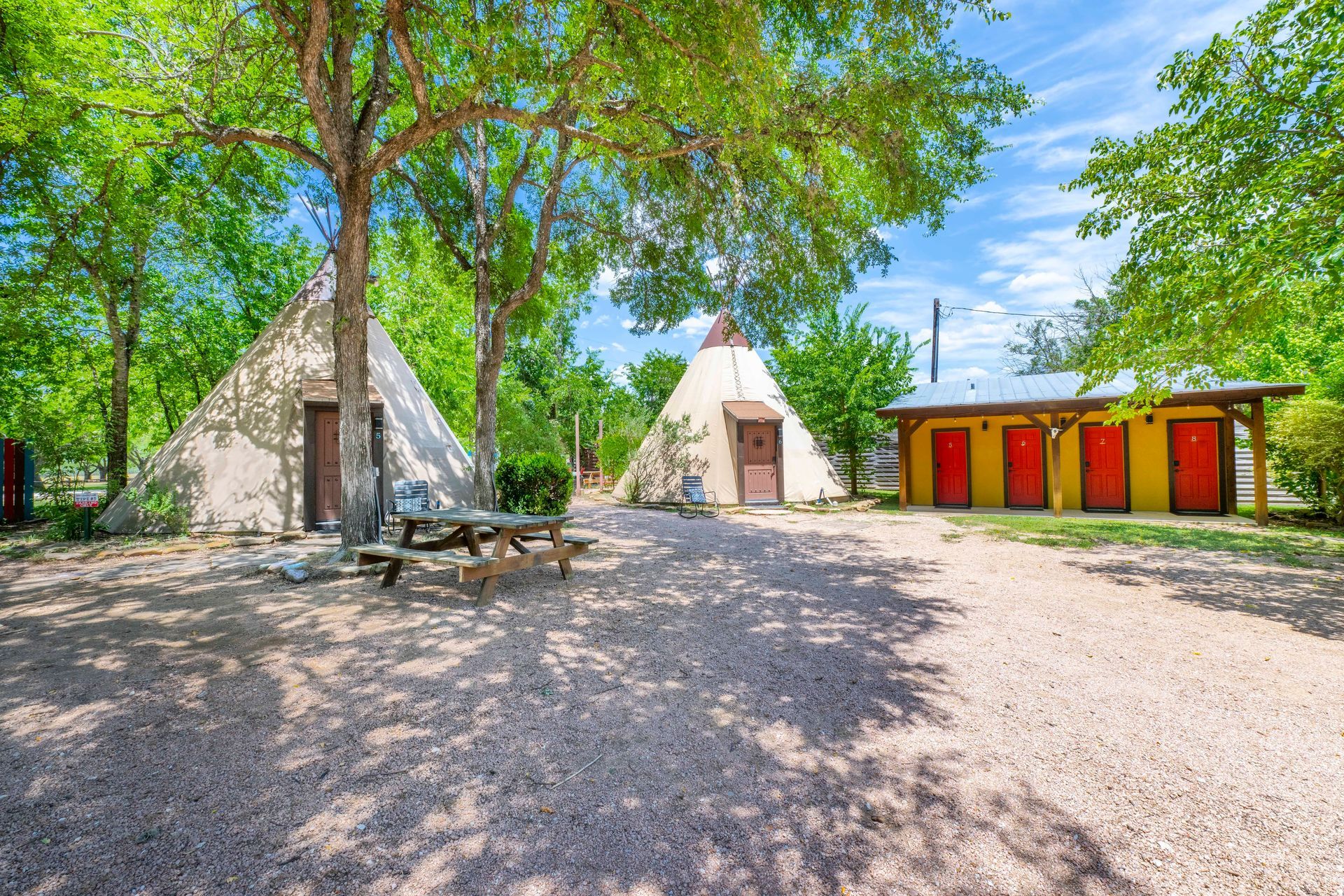 Two teepees and a red-doored bath house on gravel in a park with trees under a blue sky.