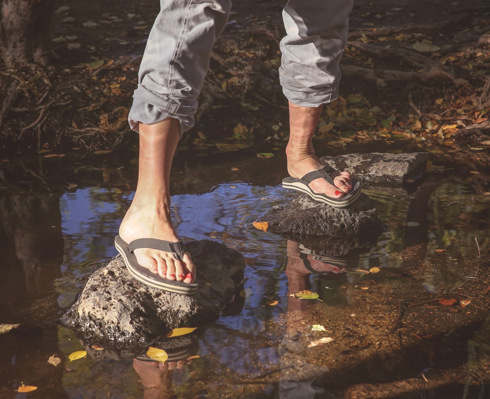 Person in flip-flops stands on a rock in a stream, pants rolled up, autumn leaves around.