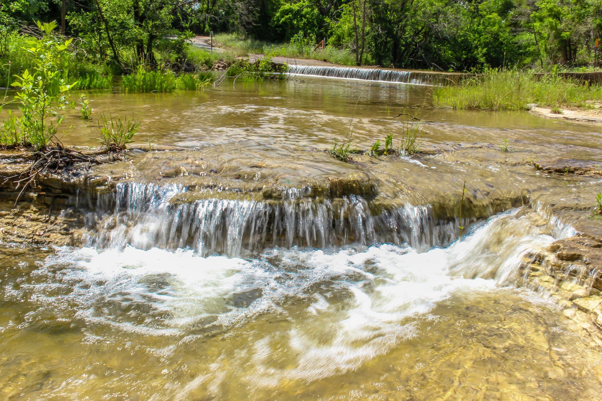 Small waterfall cascading over a rocky ledge into a shallow, flowing stream, surrounded by greenery.