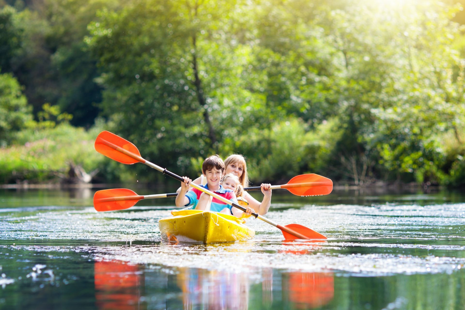 Woman and child kayaking on a calm lake, surrounded by green trees. They hold orange paddles and smile.