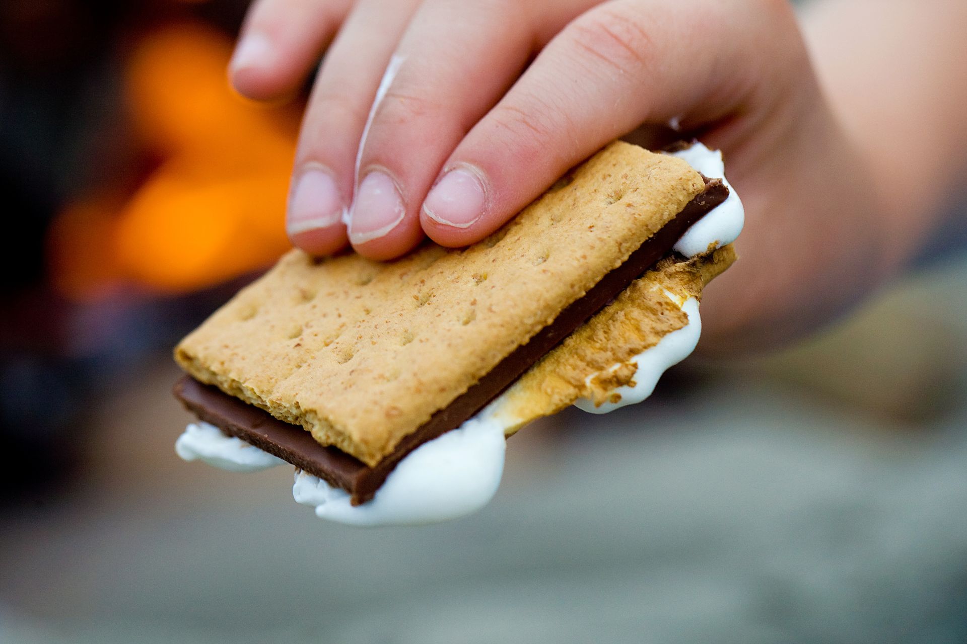 Hand holding a s'more, with melted marshmallow and chocolate between graham crackers; campfire in background.