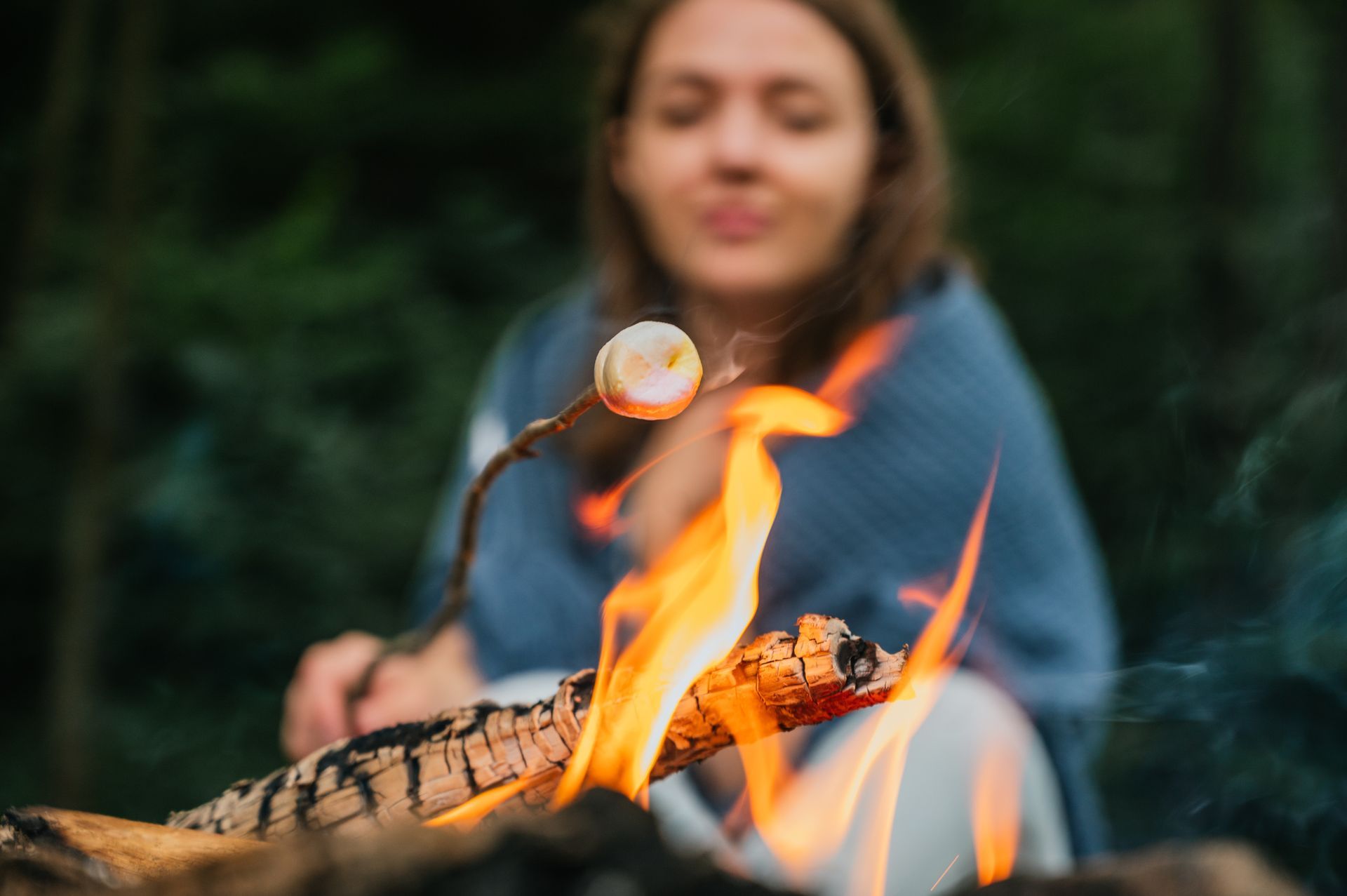 Person roasting a marshmallow over a campfire. Bright orange flames, blurred face, wrapped in a blue blanket.