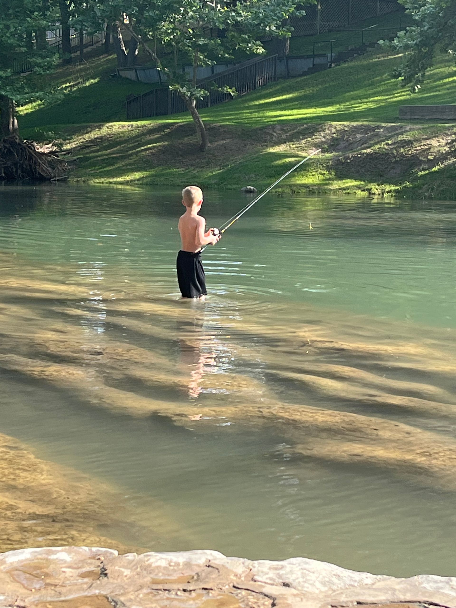 Boy fishing in a clear, shallow river. He stands waist-deep, holding a fishing rod. Green water, sunny day.