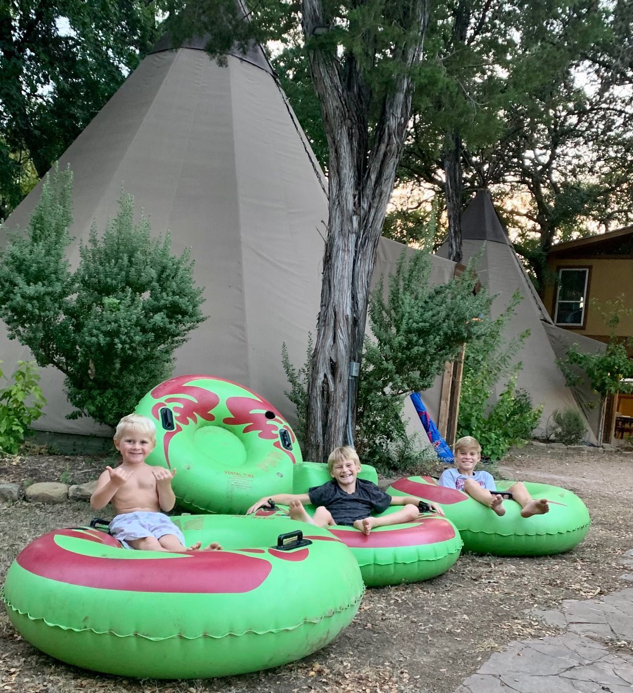 Three children in green and red inner tubes in front of tipis. They are smiling and appear ready for fun.