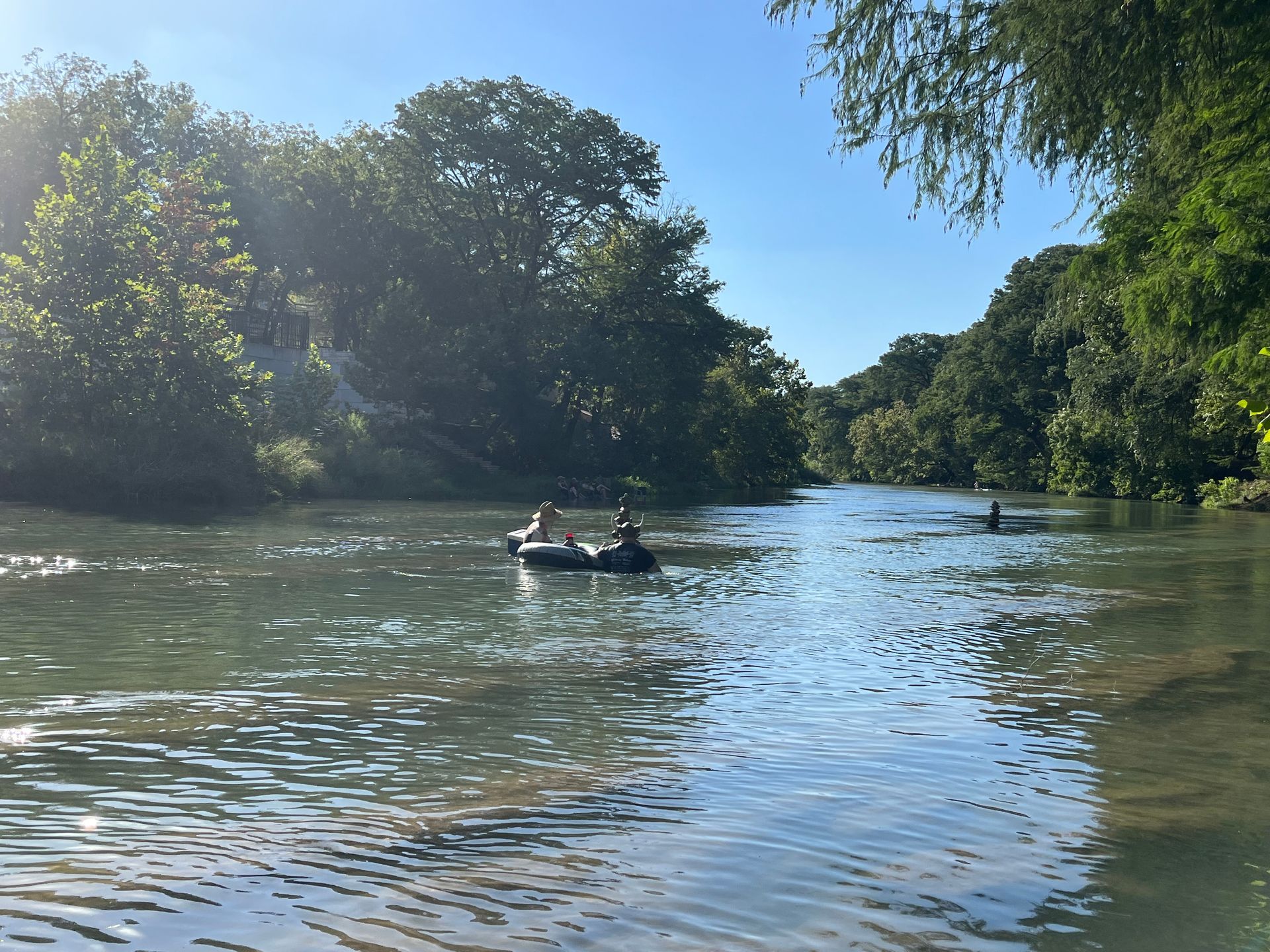 People floating on inner tubes in a river, surrounded by trees under a clear blue sky.