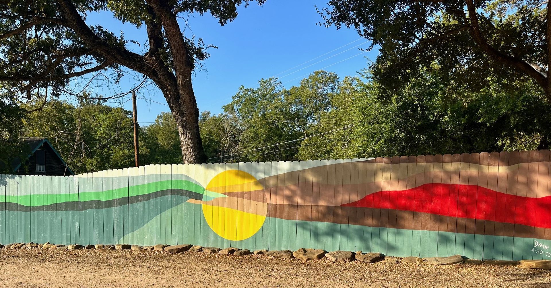 Fence with painted landscape: green hills, yellow sun, and red horizon under a blue sky.