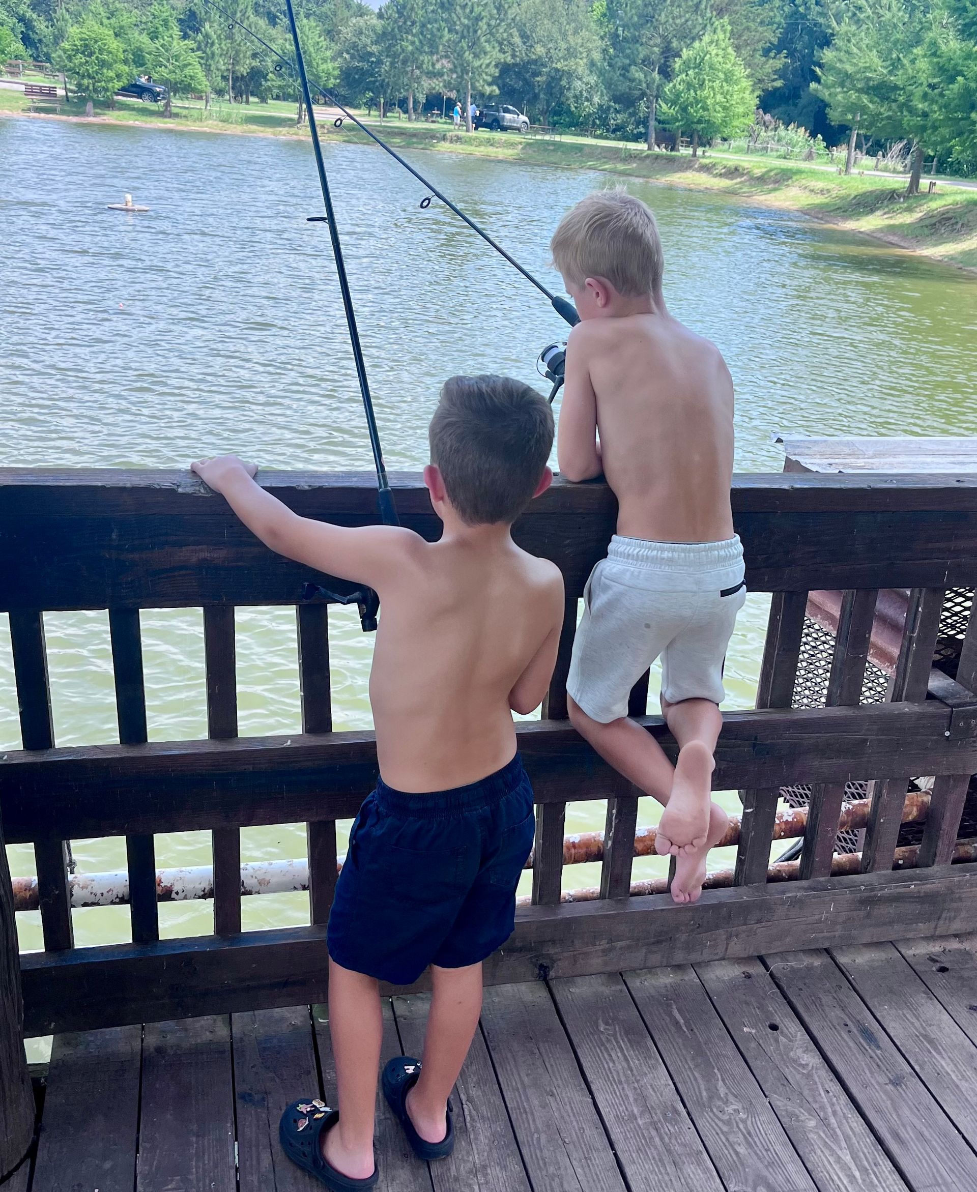 Two shirtless children fishing from a wooden dock overlooking a river.